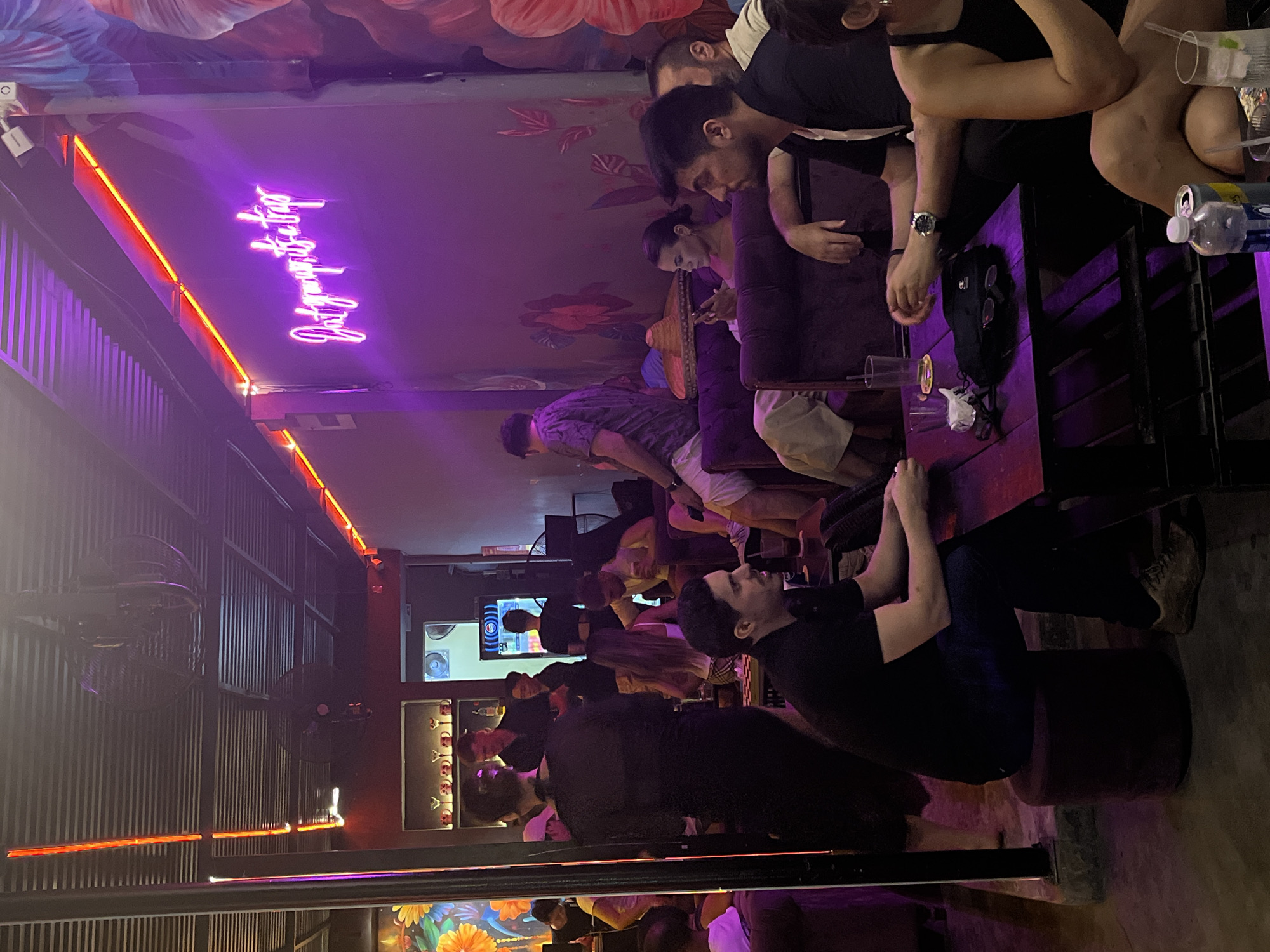 The image depicts a dimly lit indoor scene, likely a bar or restaurant in Đà Nẵng, Vietnam. Several people are visible, some seated at a long table and others standing or walking around. The interior features a colorful mural on the ceiling and wall, and neon lighting is present, including a sign that reads "Only Good Friday". Fans hang from the ceiling. The people appear to be socializing and enjoying themselves. Some are holding drinks or looking at phones. The overall atmosphere is lively and social.