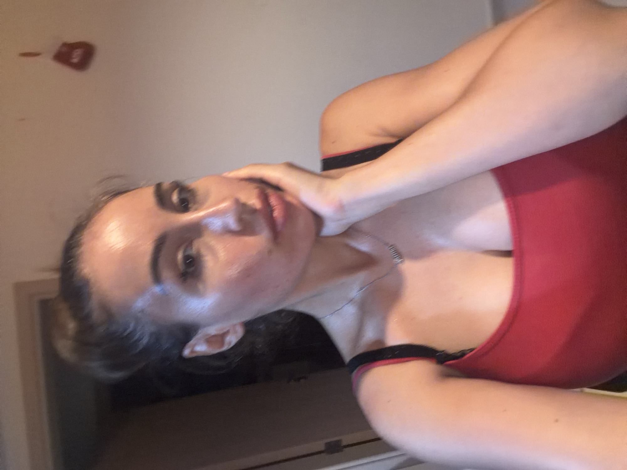 The image features a young woman with long dark hair, captured in a close-up, tilted shot. She is wearing a red top with black trim. Her left hand is touching her neck, and her gaze is directed upwards. The lighting in the image appears to be from an overhead source, creating highlights on her skin, suggesting it might be a warm environment. The background is indistinct, with a wall that has some reddish marks on it. The overall impression is that of a casual, personal photograph, possibly taken indoors. There is no visible text in the image.
