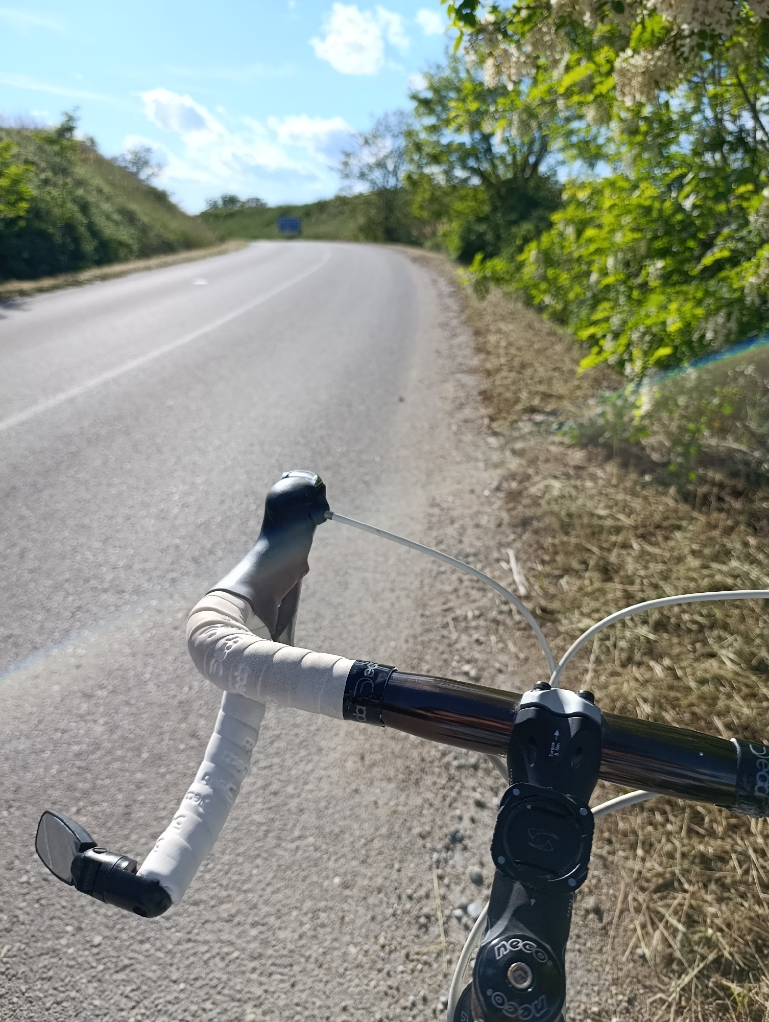 The image depicts the perspective of a cyclist riding on a paved road in what appears to be a rural or semi-rural environment. The bicycle's handlebars are in the foreground, showing white handlebar tape and a rearview mirror. The road curves gently ahead, lined on one side by vegetation, including trees and bushes. The sky is blue with scattered clouds. A sign, perhaps for traffic, is visible in the distance. The scene seems to be taken on a sunny day. The location is identified as Generalovo, Bulgaria.