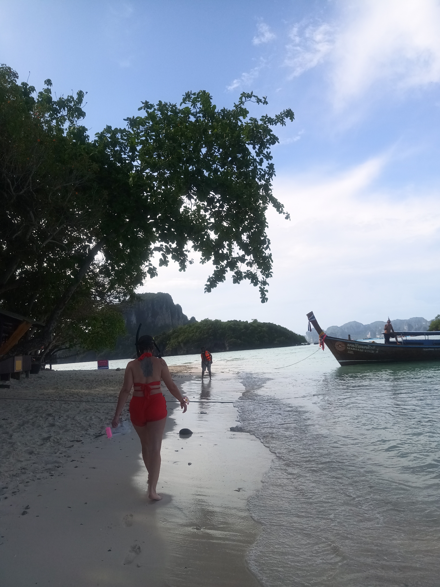 A medium shot from the back reveals a person wearing a red bathing suit and snorkeling gear walking along a sandy beach in Ao Nang, Thailand. The person is holding a pink object in their left hand. A traditional Thai longtail boat is docked in the shallow water to the right. Another person wearing an orange life vest is seen walking along the beach in the distance. Lush green trees line the shore, and rock formations rise in the background. The sky is a mix of blue and white clouds.