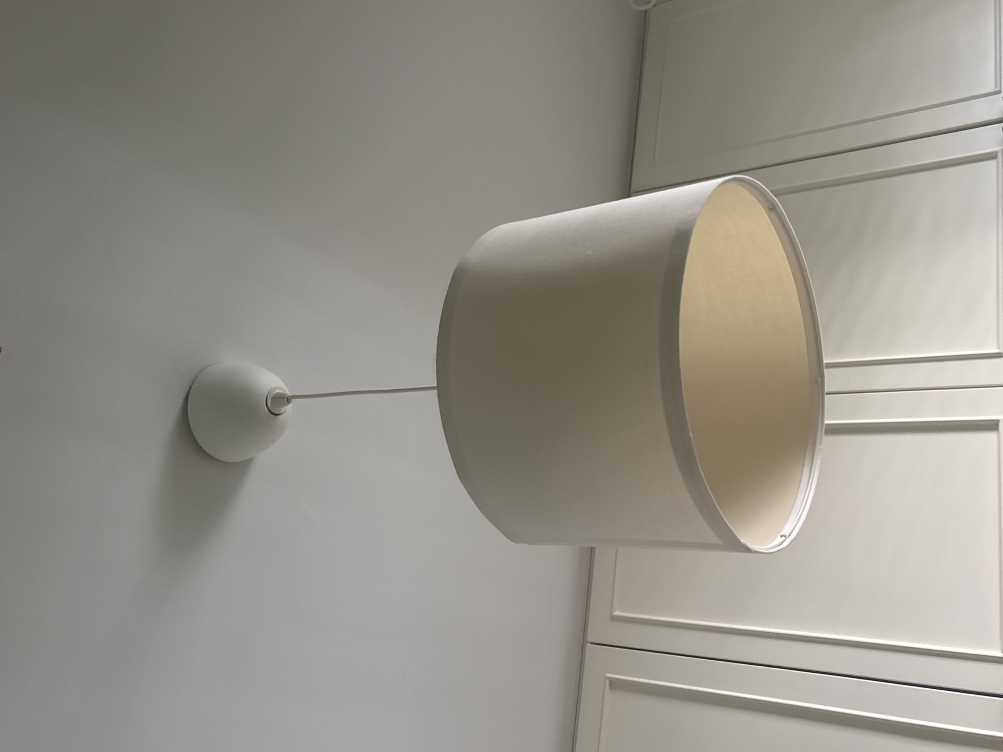 An off-white pendant lampshade hangs from a white ceiling in a room. The lampshade is cylindrical with a slightly tapered top and bottom, and a white frame. The room appears to be a kitchen or dining area, with white cabinets visible in the background. The image was taken on a bright day, and the lighting suggests it was taken indoors. No people are present in the image, and there are no visible activities or interactions. The location in the city of Lod, Israel is not discernible from the image.