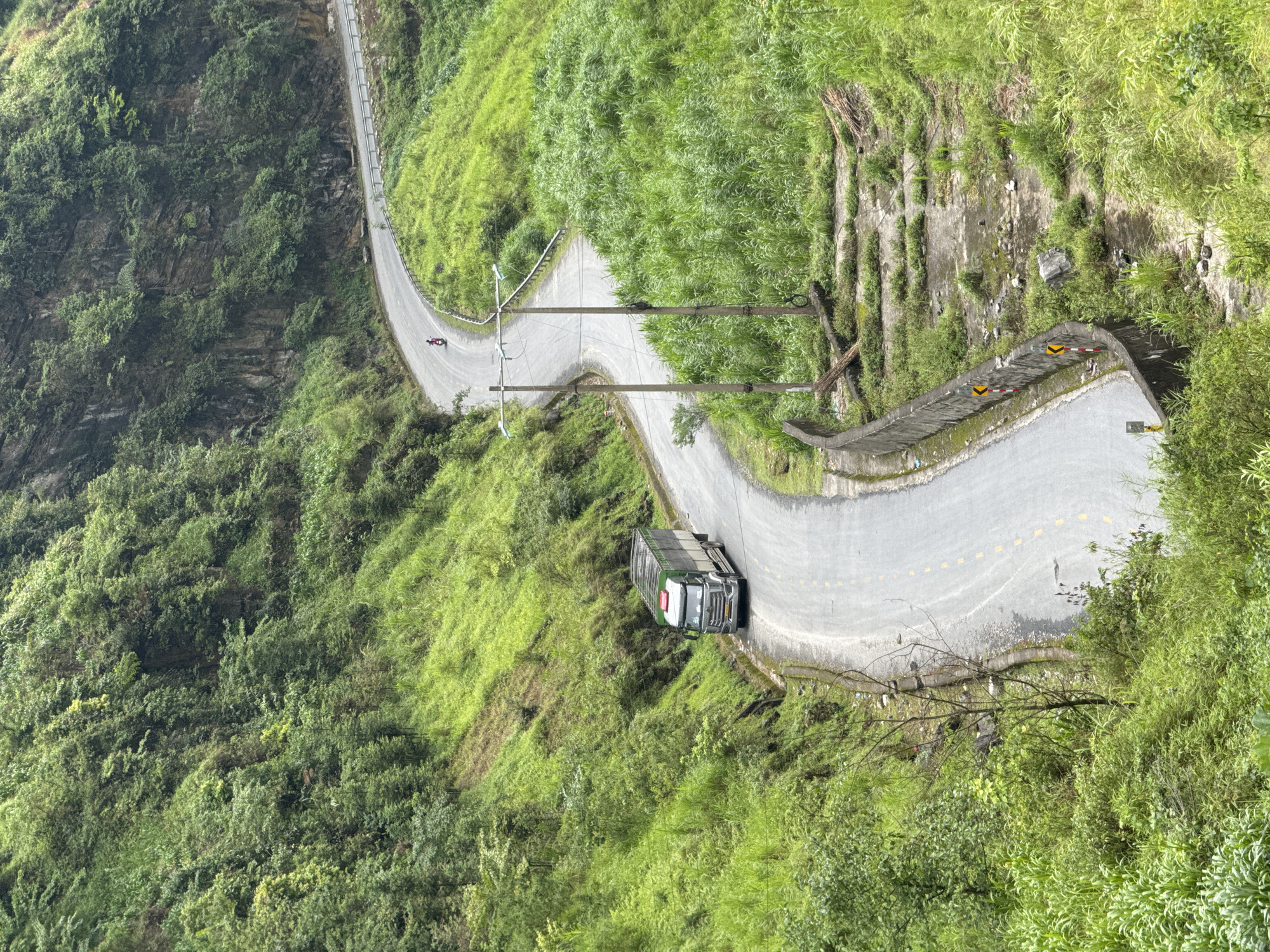 A bird's-eye view captures a winding road carved into the mountainside near the city of Yên Minh Commune, Vietnam. Lush green vegetation covers the slopes, with a grey concrete road cutting through. A truck navigates a curve in the road, while a lone figure on a motorcycle can be seen further up the path. Utility poles line the roadside, and stone retaining walls are visible on the steeper sections of the terrain.