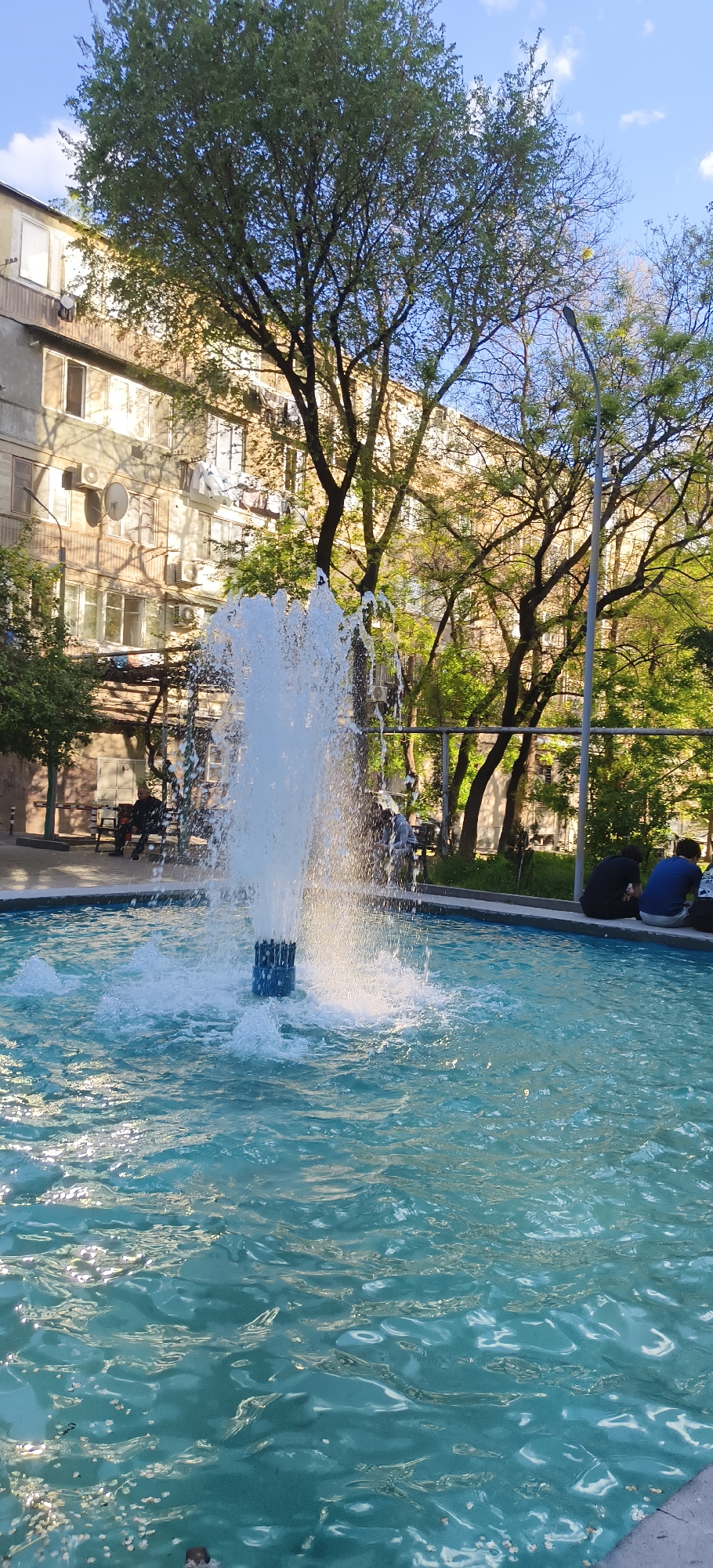A full outdoor shot shows a fountain with a turquoise-colored pool. The fountain is actively spraying water into the air. In the background are trees, a light post, a fence, benches, and a building. The building is an apartment structure. Two people are seated by the fountain’s rim. Two people are sitting on a bench in the background. The fountain is located in Yerevan, Armenia.