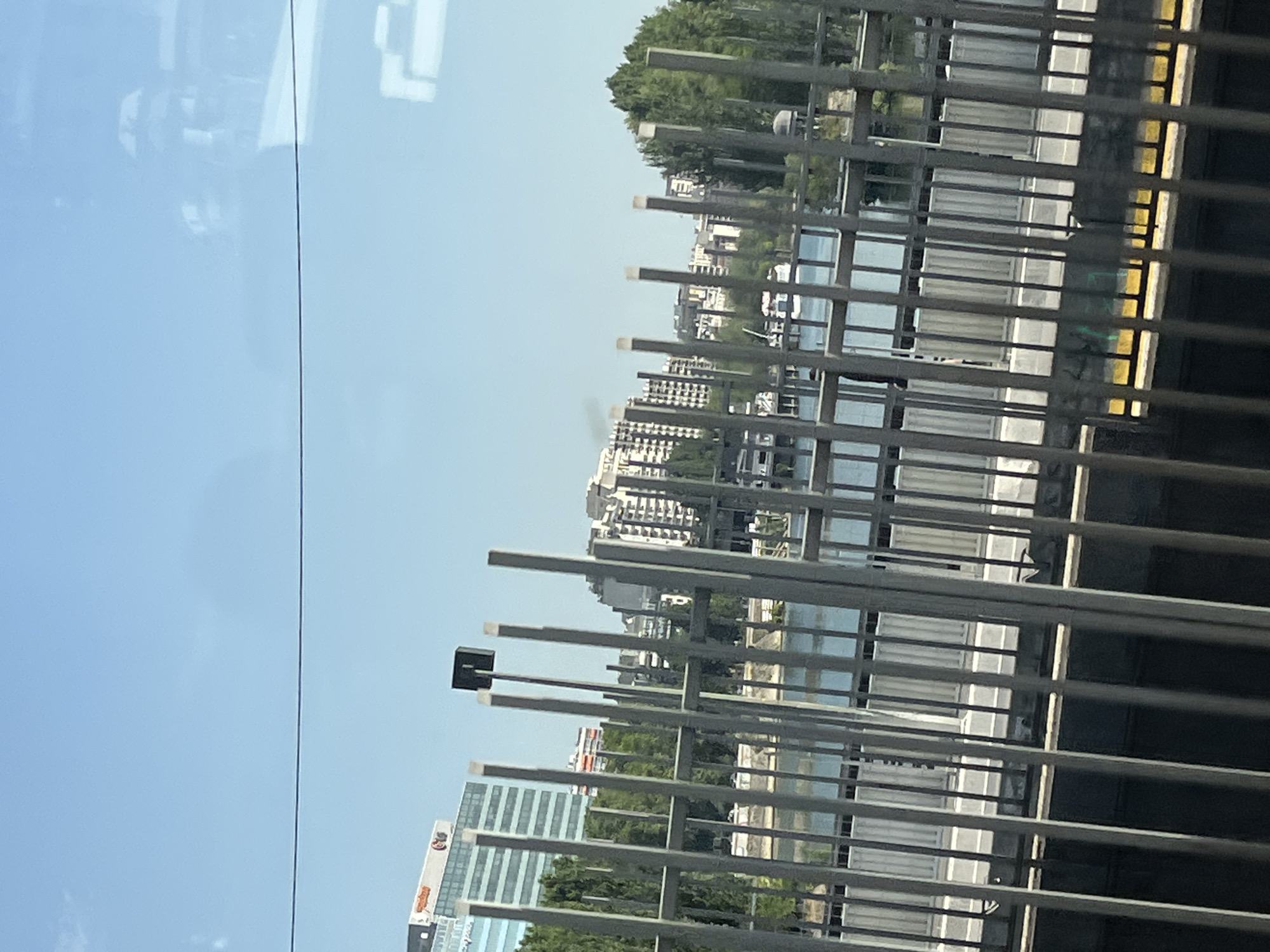 The image captures a sunny day with a clear blue sky over the city of Neuilly-sur-Seine, France. In the background, modern buildings with reflective windows are visible, some featuring logos. A body of water, likely the Seine River, separates the foreground from the buildings. The foreground is dominated by a metal fence, composed of vertical bars and horizontal railings, obscuring a portion of the view. Beyond the fence and the water, lush green trees line the riverbank. The scene appears to be taken from a slightly elevated position, possibly a bridge or an upper floor, looking across the river. There are no people or active events depicted in the image; it is a static, urban landscape.