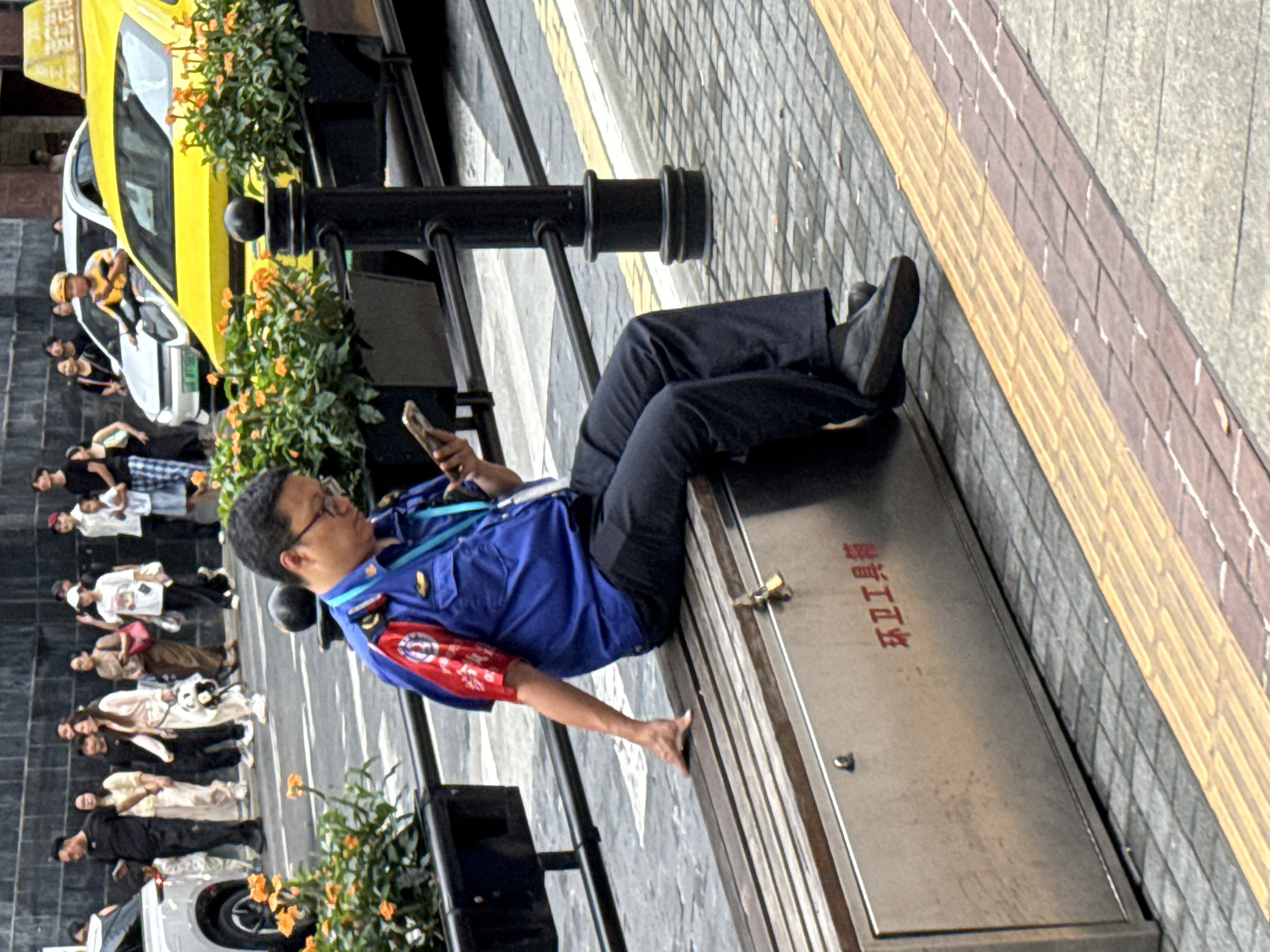 A man wearing a blue uniform with red accents is sitting on a wooden bench in Yuzhong District, China. He is holding a mobile phone and appears to be looking at it. The bench is positioned next to a metal cabinet with Chinese characters that read "环卫工具箱" (Environmental Sanitation Tool Box). Behind him, a yellow taxi and other vehicles are visible on a street. The environment suggests an urban setting with pedestrian walkways and a street. The man's uniform implies he may be an employee of the sanitation department.
