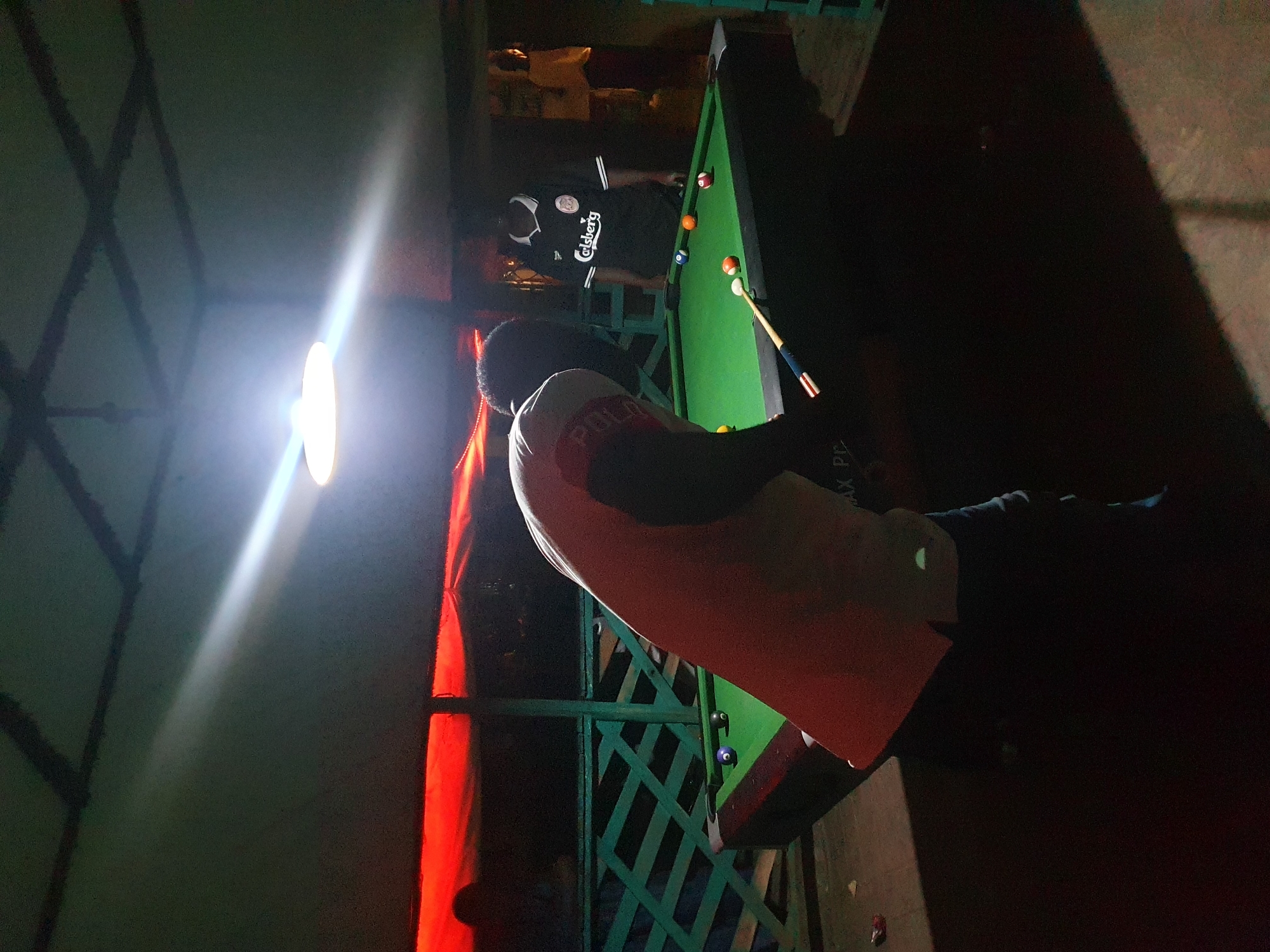 The image depicts a dimly lit scene in what appears to be an outdoor or semi-outdoor area. A man wearing a light-colored shirt with dark sleeves and the word "POLAR" on it is leaning over a pool table. The pool table is green, with several pool balls visible. A pool cue rests on the table near some of the balls. Another man is visible in the background, wearing a black Carlsberg shirt. The lighting is poor, with a bright light source in the upper-left corner casting harsh shadows. The background includes a green lattice-like structure and a wall with red lighting. It is not possible to confirm the location in Lokoja, Nigeria, based solely on the image. The main activity shown is the playing of pool.