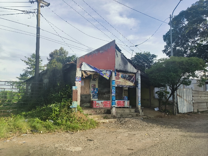 A small, weathered, red-roofed building with a shopfront is located at the coordinates -6.753, 107.444 in the city of Cikalong, Indonesia. The shopfront is covered in advertising posters, and there are signs in both Indonesian and English. The building is located on the edge of a dirt road, and there are trees and bushes in the foreground. There is a utility pole with multiple powerlines running to it, and another utility pole with streetlights. The building is facing a small, dirt road, and there is a fence made of iron bars to the left of the building. To the right of the building is a larger, unpainted, metal building. There are trees in the background, with a blue, clear sky above. There are no people in the scene. The scene is likely from a rural area.