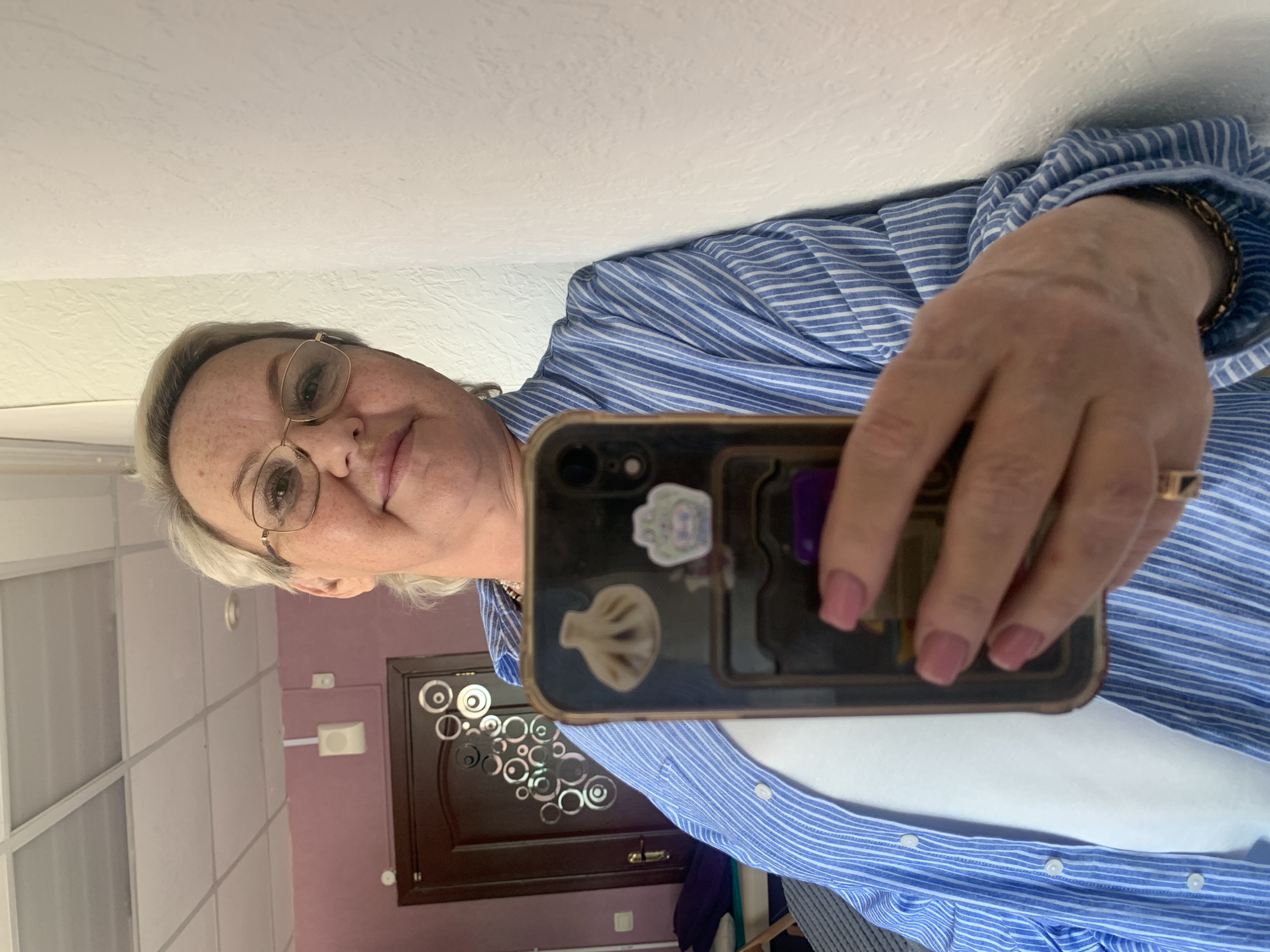 A selfie of a woman with short gray hair and glasses, wearing a blue and white striped shirt over a white top. She is holding a smartphone with stickers on the case. Behind her is a doorway with decorative circles. The room appears to have light purple walls and a white ceiling. There is no explicit information in the image to confirm that the location is Chaikovsky, Russia.