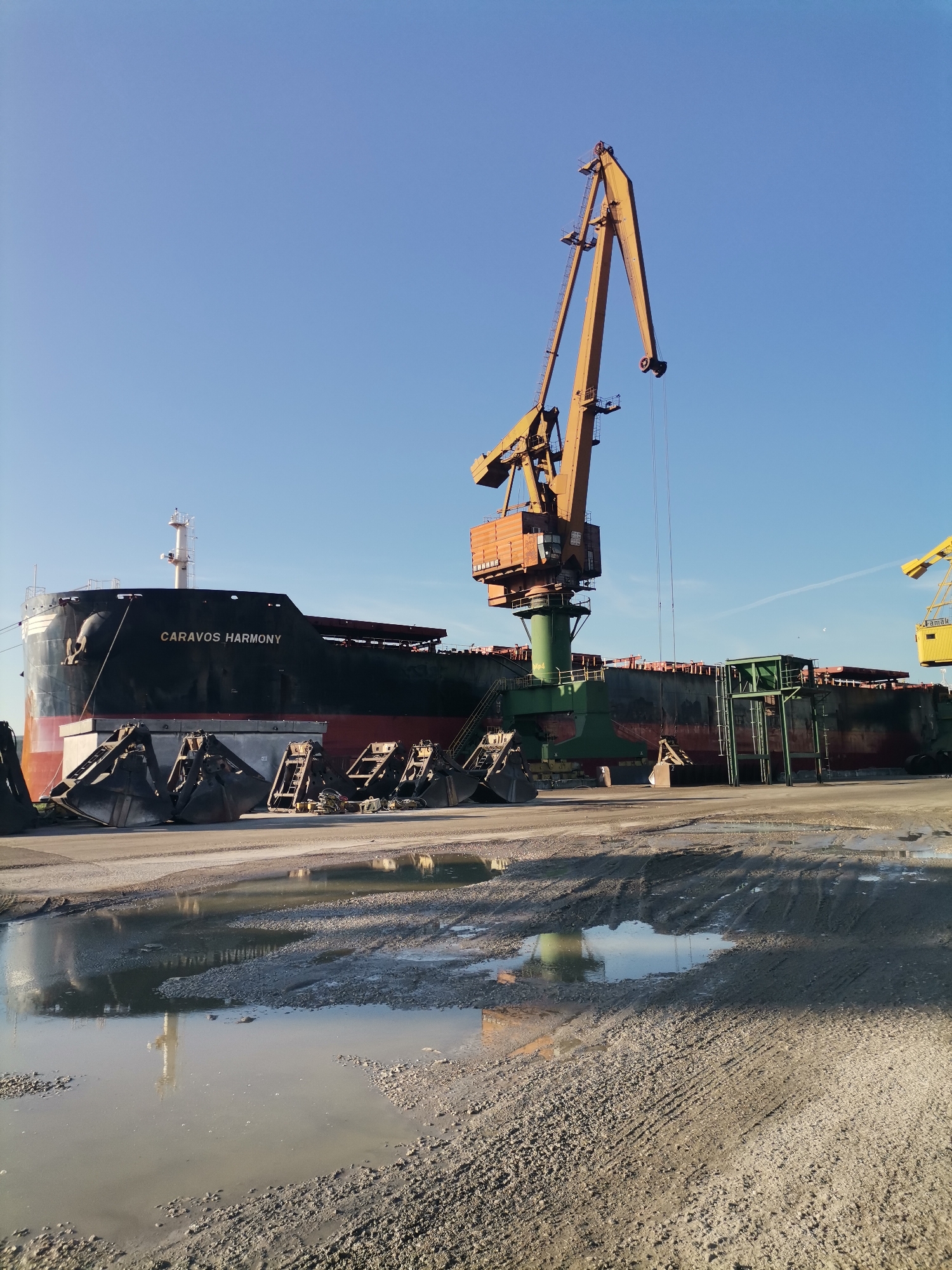 The image shows a large cargo ship, named "CARAVOS HARMONY", docked at a port in Ezerovo, Bulgaria. The ship is black and red, and it is flanked by cranes used for loading and unloading cargo. There is a large orange crane to the right of the ship. In front of the ship, there are several large metal clamshell buckets and machinery, along with puddles on a muddy, gravel-covered surface. The sky is blue and clear.