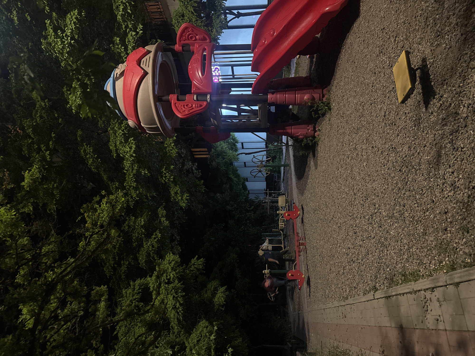 The image shows a playground at night. The playground equipment includes a red slide, a rocket-shaped structure with red and beige accents, a yellow square panel, and a seesaw. The ground is covered with small gravel. There are trees bordering the playground. In the background, light from an open sign is visible. The location is in Yerevan, Armenia.