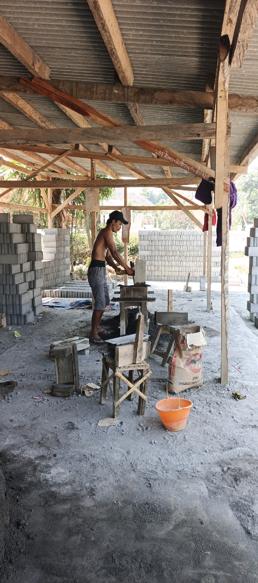 A man is working in an open-air shed, making bricks in Kasiyan, Indonesia. He is wearing a black baseball cap, grey shorts, and no shirt, and he is using a hammer to strike a brick mold. Stacks of gray bricks are visible in the background, as well as a bag of cement and an orange bucket. The shed has a corrugated metal roof supported by wooden beams, and the ground is covered with dirt and small stones. There are trees and foliage visible in the distance.