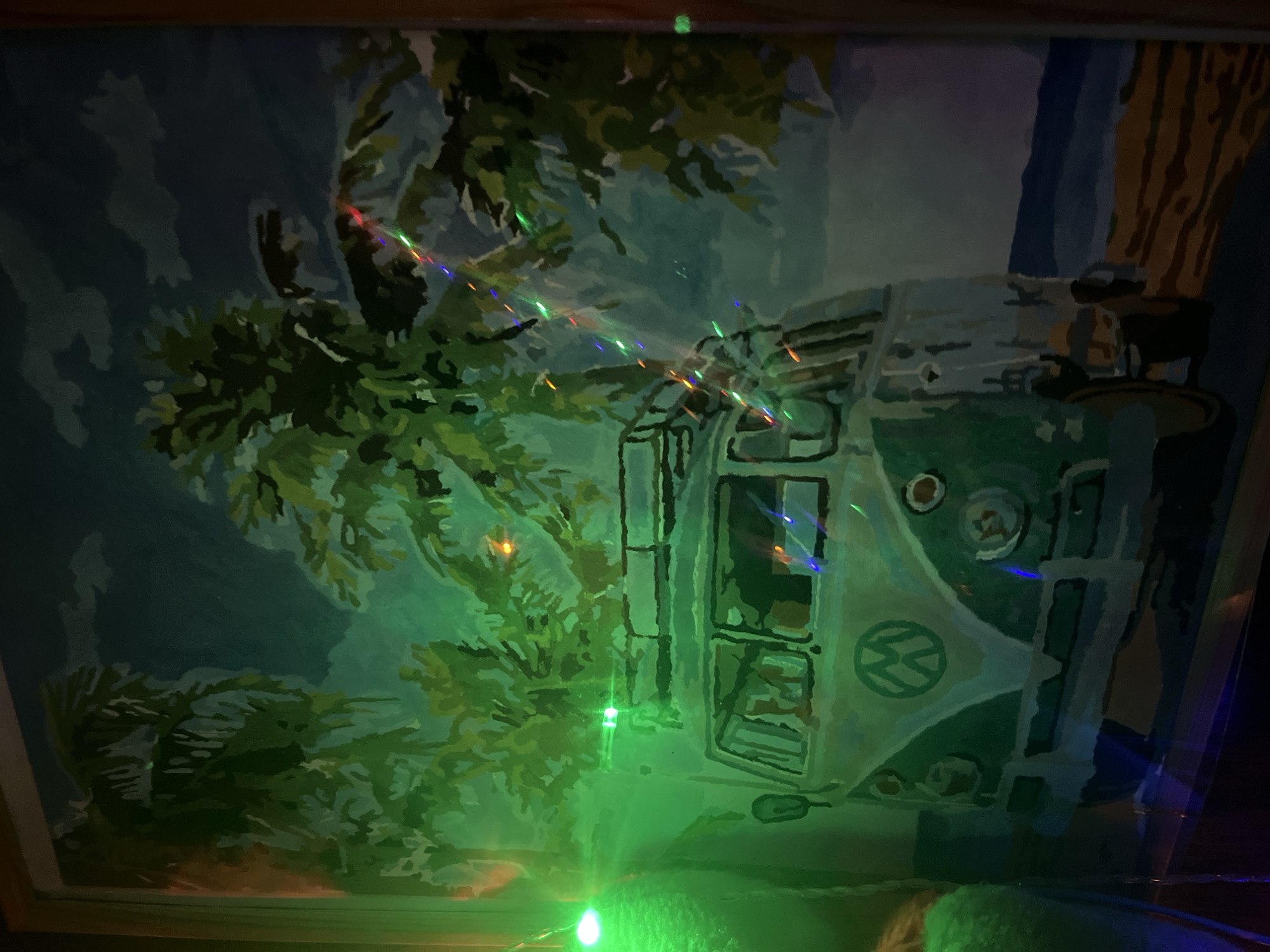 The image depicts a painting of a vintage Volkswagen van parked on a beach with palm trees in the background. The painting is illuminated by green and multi-colored string lights, which cast reflections on the surface. The scene is framed by what appears to be a wooden border. There is no discernible information within the image to indicate its location in the city of Kazan, Russia.