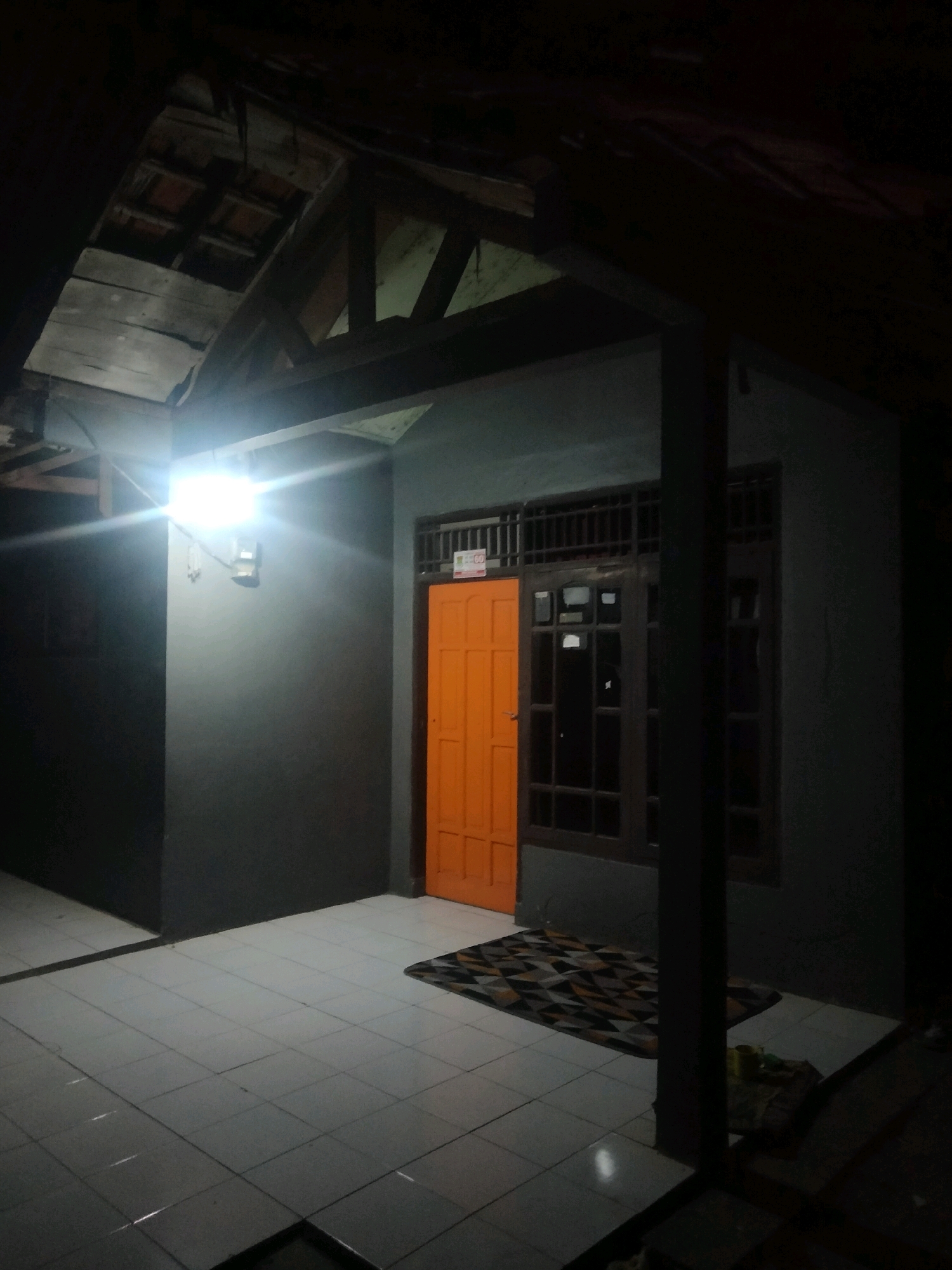 The image depicts the exterior of a building at night, illuminated by a bright light source. The building appears to be a residential structure, with a tiled floor in front. An orange door is visible, along with a window. The roof structure is partially exposed, revealing wooden beams and tiles. A mat lies on the tiled floor in front of the door. The overall environment suggests a simple, potentially rural or suburban setting in Suka Sari, Indonesia. There are no people visible in the image, and no apparent criminal activity is depicted.