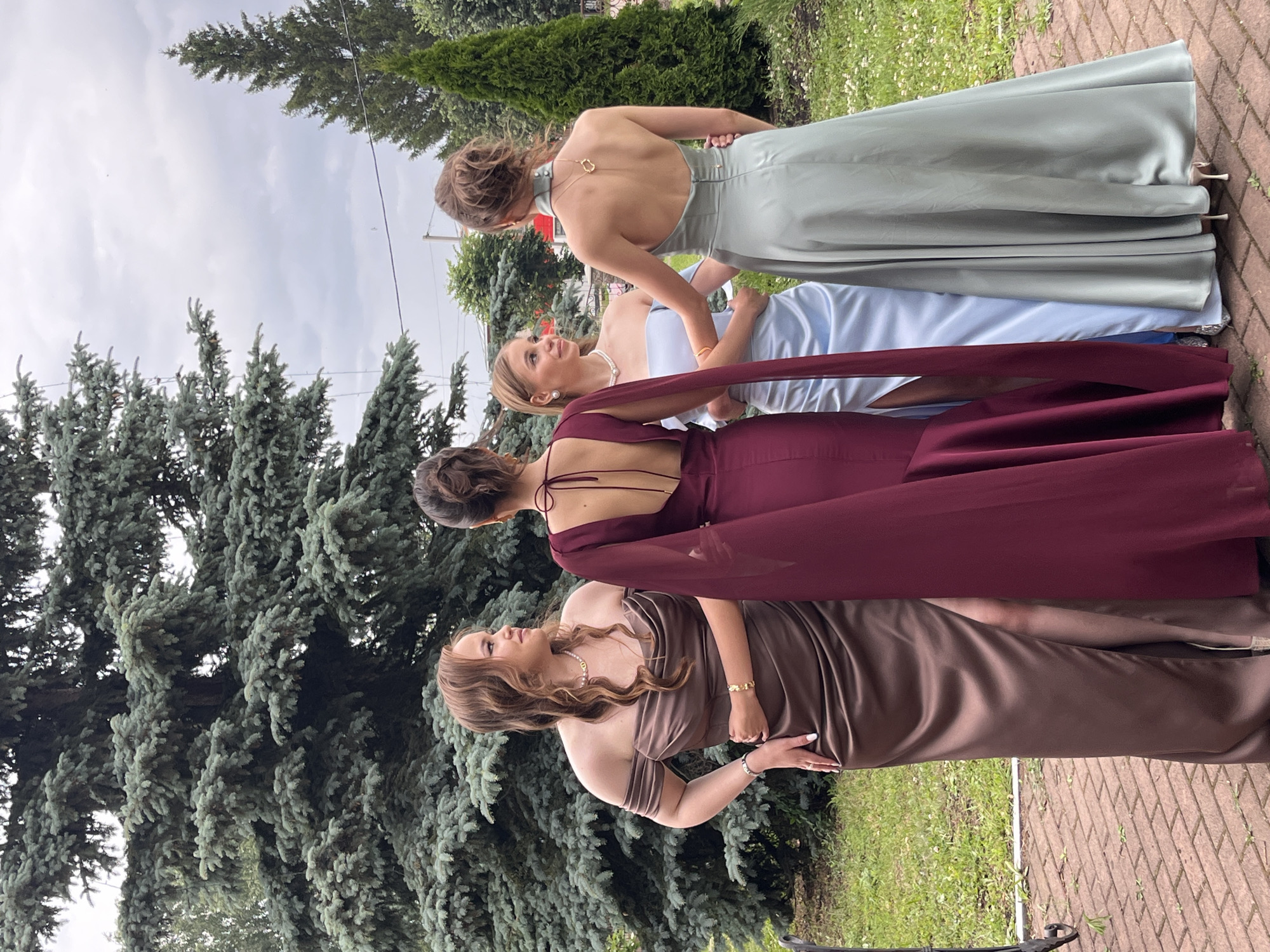 Four young women are standing in a circle, facing away from the camera, in what appears to be a garden setting in Starobaltachevo, Russia. They are dressed in formal, floor-length gowns. The woman on the far left wears a brown off-the-shoulder dress with a slit up the leg. Next to her, a woman in a deep red dress with a cape-like overlay. The third woman from the left is in a light blue off-the-shoulder dress. The woman on the far right is in a pale green halter dress. They are positioned in front of a large evergreen tree, with a smaller evergreen tree and other foliage visible in the background. The ground is paved with red bricks. The sky is overcast.