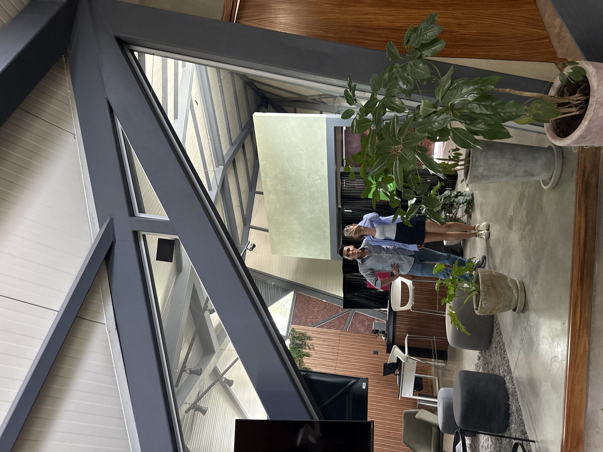 The image shows a modern interior, possibly in San Bernardino Tlaxcalancingo, Mexico. The architecture features a triangular frame structure, predominantly gray, with glass panes. The interior design includes wooden walls with geometric patterns and off-white panels. A couple stands within the interior space. Various objects like chairs, stools, and a TV monitor are visible. Plants in pots are also present.