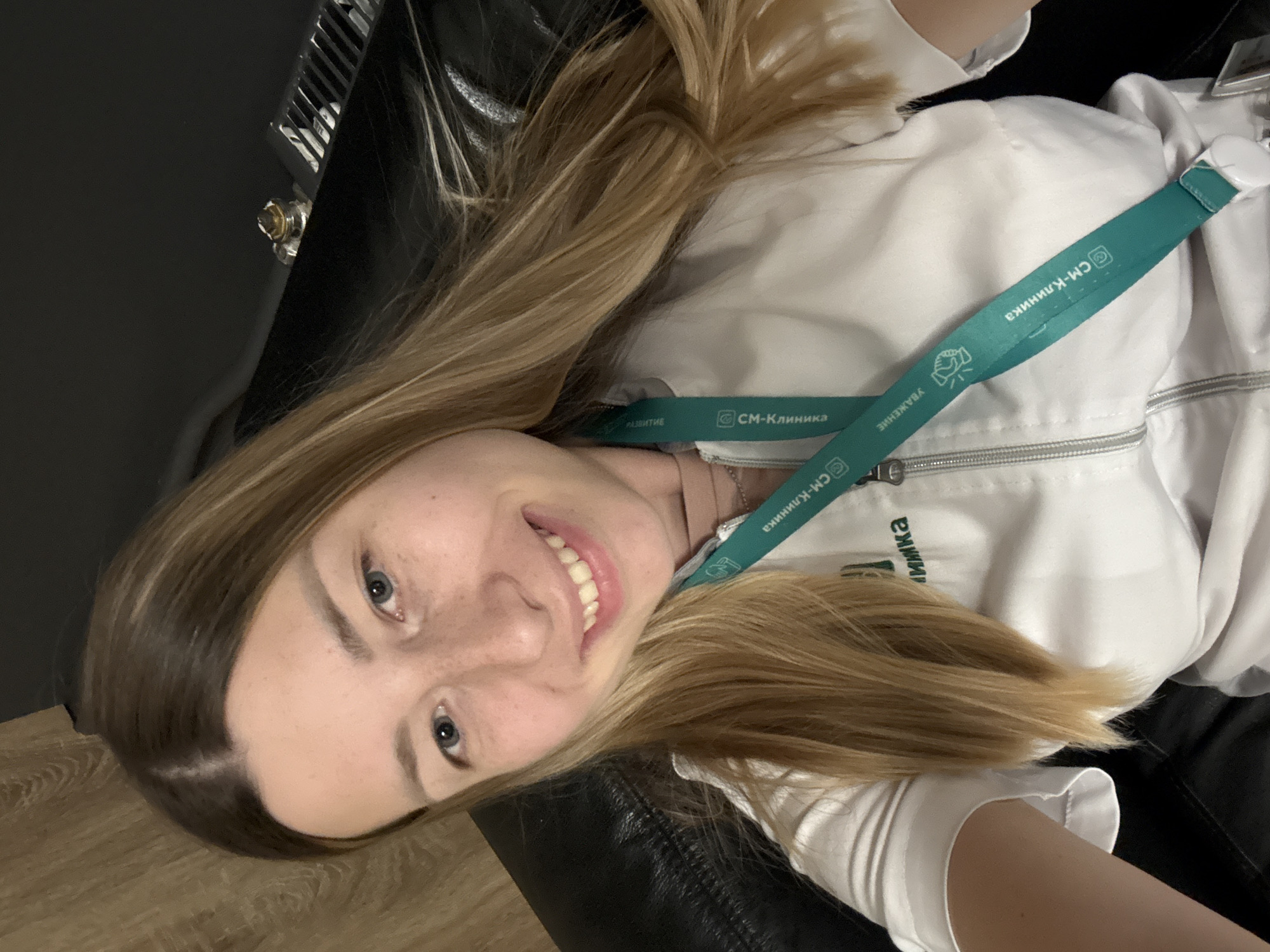 A close-up, selfie-style photograph shows a young woman with shoulder-length blonde hair, fair skin with freckles, and blue eyes. She is smiling and looking directly at the camera. She is wearing a white lab coat or uniform over a light-colored top, and a teal lanyard with the text "СМ-КЛИНИКА" (SM-CLINICA) is visible around her neck. The background is dark and out of focus, appearing to be an indoor setting, possibly a room with a black leather couch or chair and a wooden wall. There is no identifiable information to indicate the location within Moscow, Russia.
