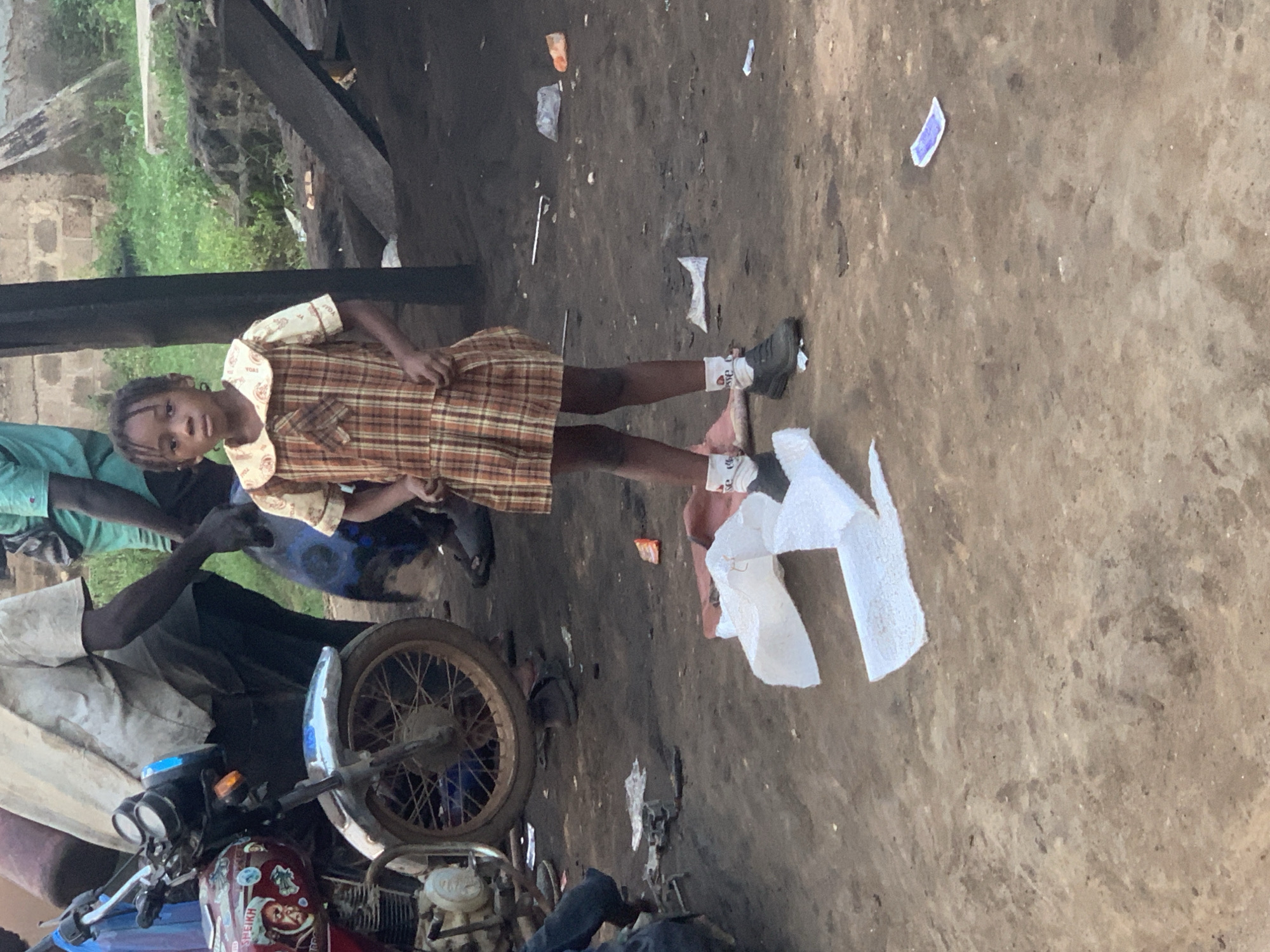 A young Black girl stands in a dusty outdoor area. She is wearing a plaid dress and white socks with dark shoes. The girl has dark hair pulled back and is looking towards the camera. Behind her, a person in a light-colored shirt and dark pants is visible next to a motorcycle. Further in the background, other people and greenery can be seen. The ground is covered in dirt and debris, with some white material and a purple item scattered on the ground. The scene appears to be in a marketplace or workshop area. This image was taken in the city of Shagamu, Nigeria.