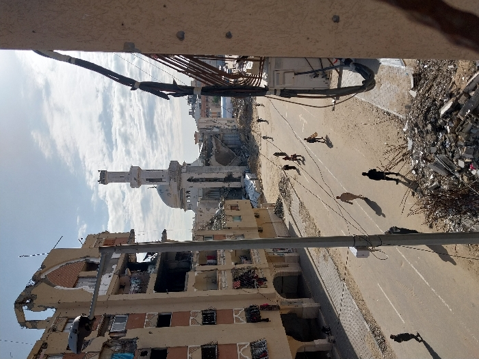 A devastated cityscape features buildings riddled with holes and rubble, suggesting heavy conflict. A partially destroyed mosque with a minaret stands amidst the destruction. Several people are walking on a road that runs through the scene. Electrical wires and concrete structures frame the view, adding to the feeling of urban decay.