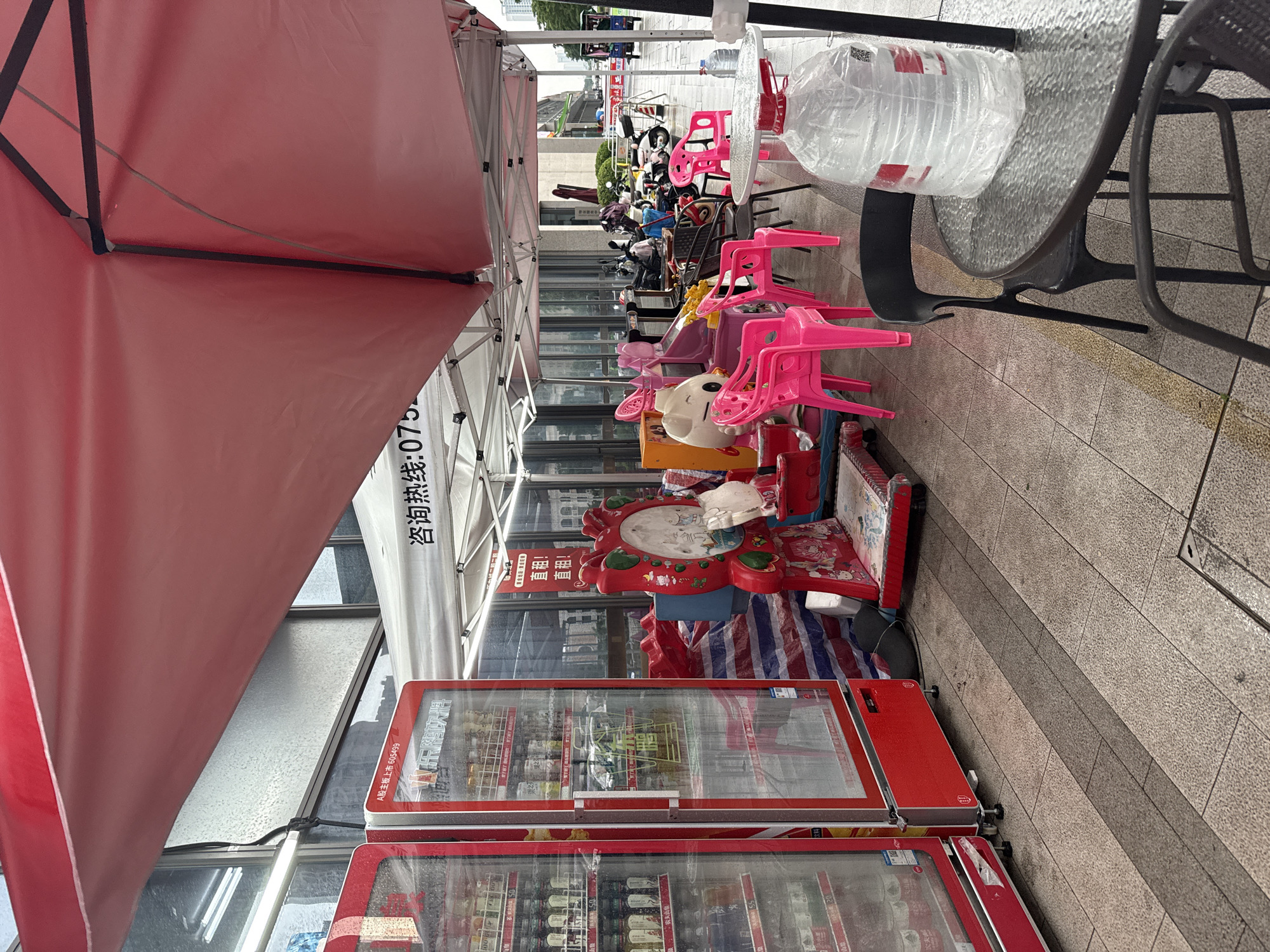 The image depicts an outdoor scene in Huizhou, China, under a red awning. In the foreground, a glass-top table with black chairs is visible. A large, clear plastic-wrapped bottle of water sits on the table. To the left, two red vending machines are filled with beverages. Behind the vending machines, a tent is set up. Several colorful children's rides, including a red one with a mirror and a white character, are present. Pink plastic chairs are scattered around the area. In the background, various motorcycles and scooters are parked. The ground is paved with grey tiles, and the overall environment appears to be a street or sidewalk area.