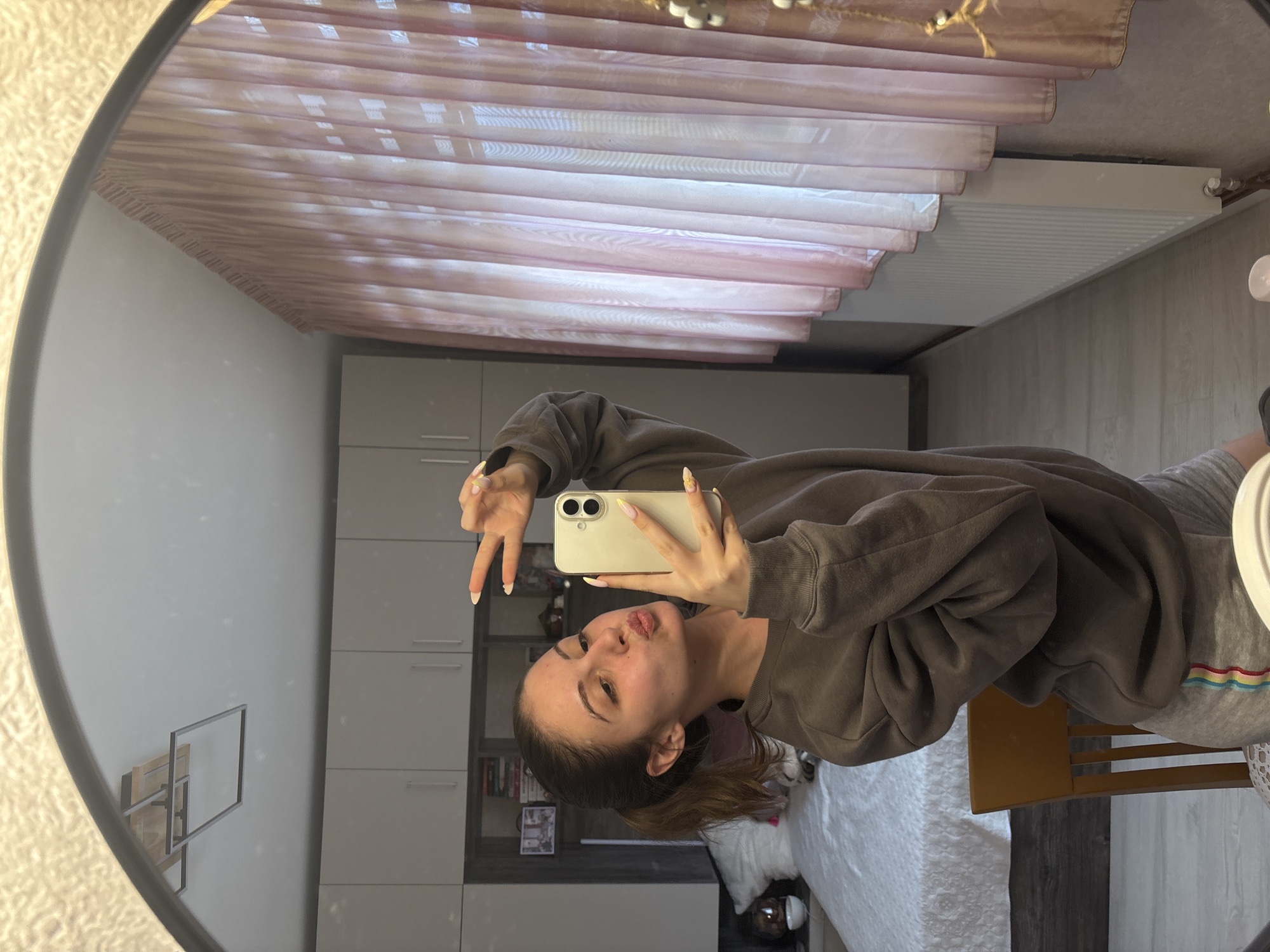 The image depicts a young woman in a room reflected in a circular mirror. She is holding a phone, presumably taking a selfie, and making a "peace" sign with one hand while puckering her lips. She is wearing a brown sweatshirt and grey sweatpants. The background includes a bed with a white comforter, a white radiator, and some furniture, potentially wardrobes or cabinets. Pink curtains are visible above. The location is not specified in the image, therefore Bulgarovo, Bulgaria cannot be confirmed or denied.