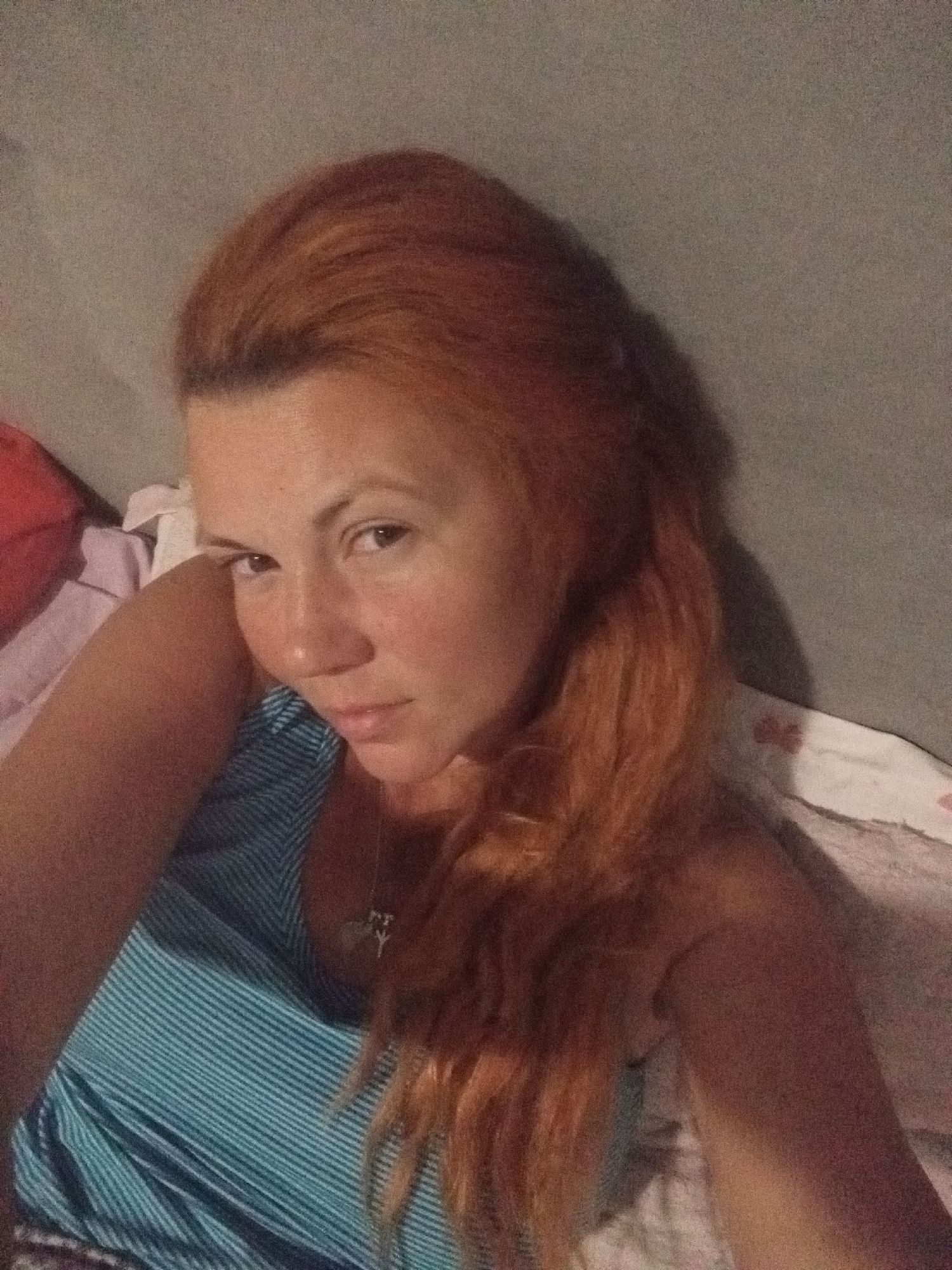A young woman with long, reddish-brown hair, styled to one side, lies on her side and looks towards the camera. She is wearing a blue and white striped tank top and a silver necklace with a cross pendant. Her skin appears tanned and slightly flushed. She has a neutral expression. The background is indistinct, but appears to be a bed with pillows and blankets. The lighting is dim, suggesting it could be nighttime or indoors with low light. The overall impression is a casual, personal photograph, likely a selfie taken by the subject. There is no visible text.