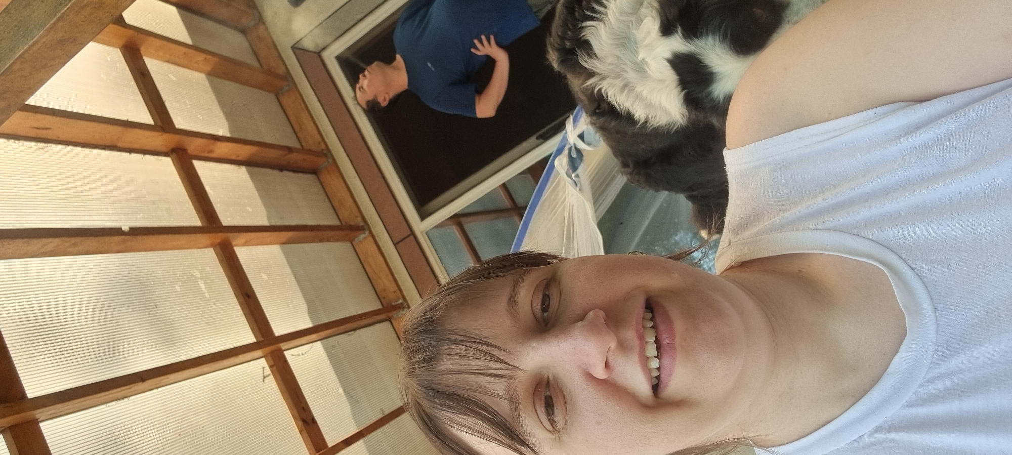 The photograph presents an indoor scene with three individuals—a woman, a man, and a dog—along with the architectural structure of a room with a grid-like ceiling. The woman, positioned in the foreground, wears a white T-shirt and looks directly at the camera with a slight smile. Behind her and above, a man in a blue shirt is visible through an open window-like structure. A black and white dog partially occupies the upper right corner of the image. The location appears to be indoors, possibly a sunroom or enclosed patio, characterized by a wood-framed, grid-patterned ceiling that filters sunlight. While the specific city of Dunnae-myeon, South Korea, is mentioned, there is no specific visual information in the image that confirms or denies that geographic location. The scene is illuminated by natural light, and the overall composition suggests an informal, candid moment.