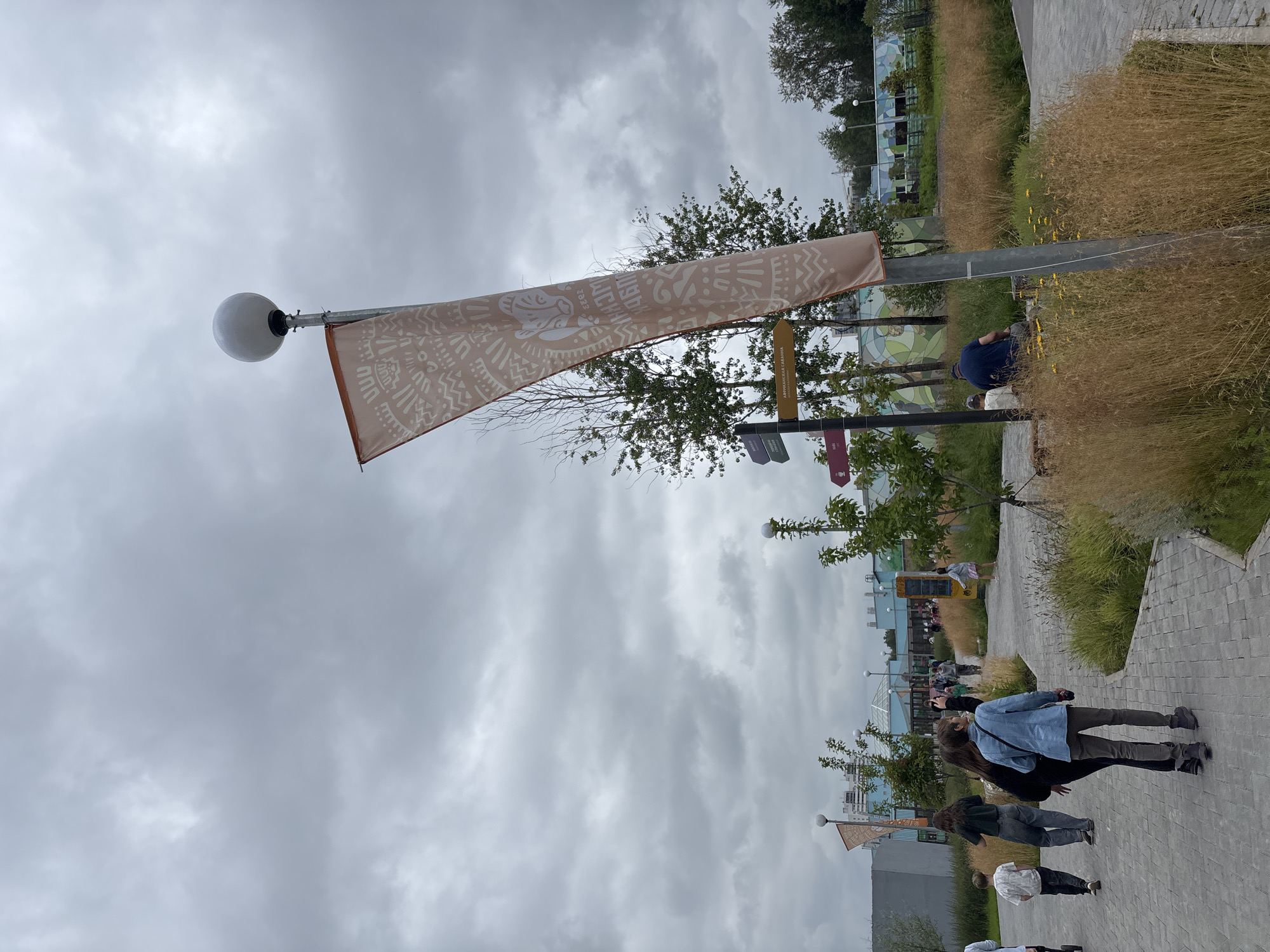 An outdoor, overcast scene depicts a paved pathway winding through a park-like area with dry grasses and trees. A tall metal pole with a spherical light fixture stands prominently, adorned with a light tan flag featuring a tribal pattern. Several people are walking on the pathway, some in groups and some individually. In the background, buildings with colorful murals are visible. A signpost with directional arrows, one reading "АФРИКАНСКАЯ САВАННА," is also present. The overall impression is that of a public recreational space on a cloudy day.