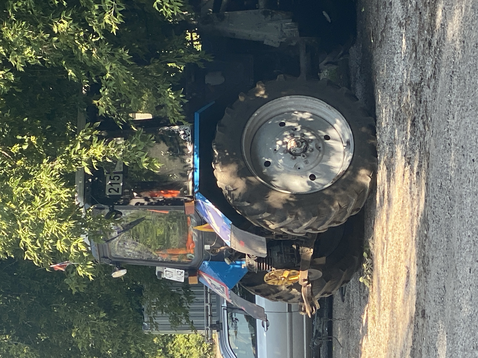 A blue and silver tractor with large rear tires is parked on a gravel surface, partially obscured by green foliage from a tree. A silver car is visible to the left of the tractor. The tractor has a license plate that reads "4257 KK 42". The number "215" is displayed on the front of the tractor's cab. A small flag is visible on top of the tractor. The scene appears to be outdoors in an area that could be a rural or industrial part of Novokuznetsk, Russia, judging by the gravel, car, and utilitarian nature of the tractor. No people are visible in the image.