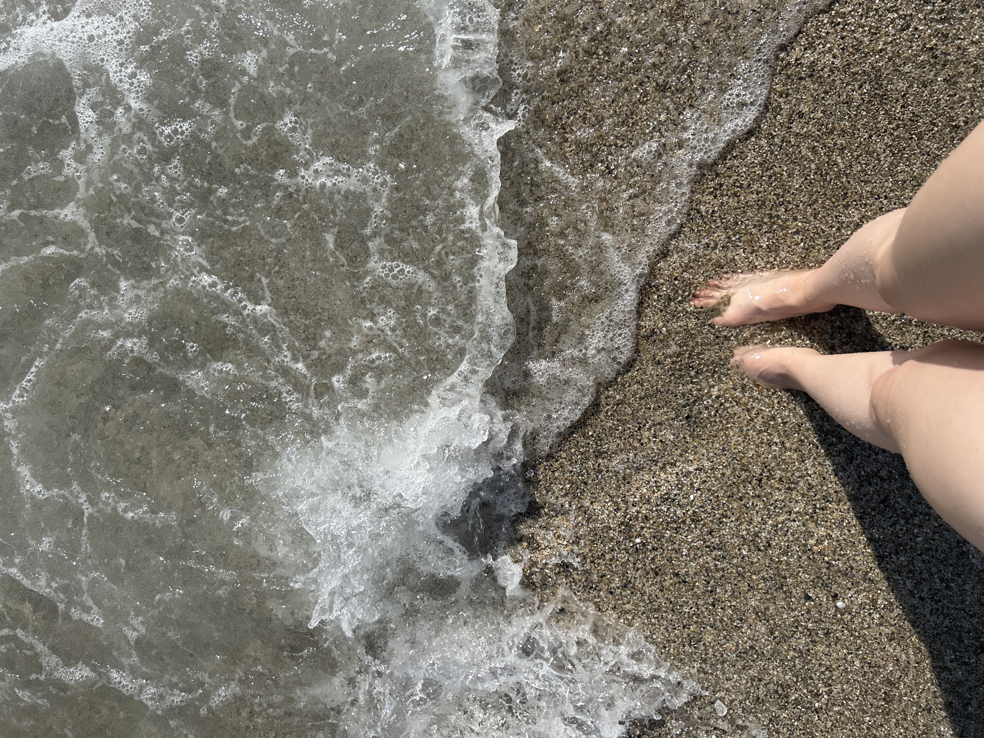 The image shows a beach in an unknown city. The photograph, taken from an overhead perspective, captures the meeting point of water and sand. On the right side of the frame, two legs and feet are visible on the sand, suggesting a person is present. The sand appears coarse and textured. The water is a gray hue with white foam and bubbles, indicating movement.