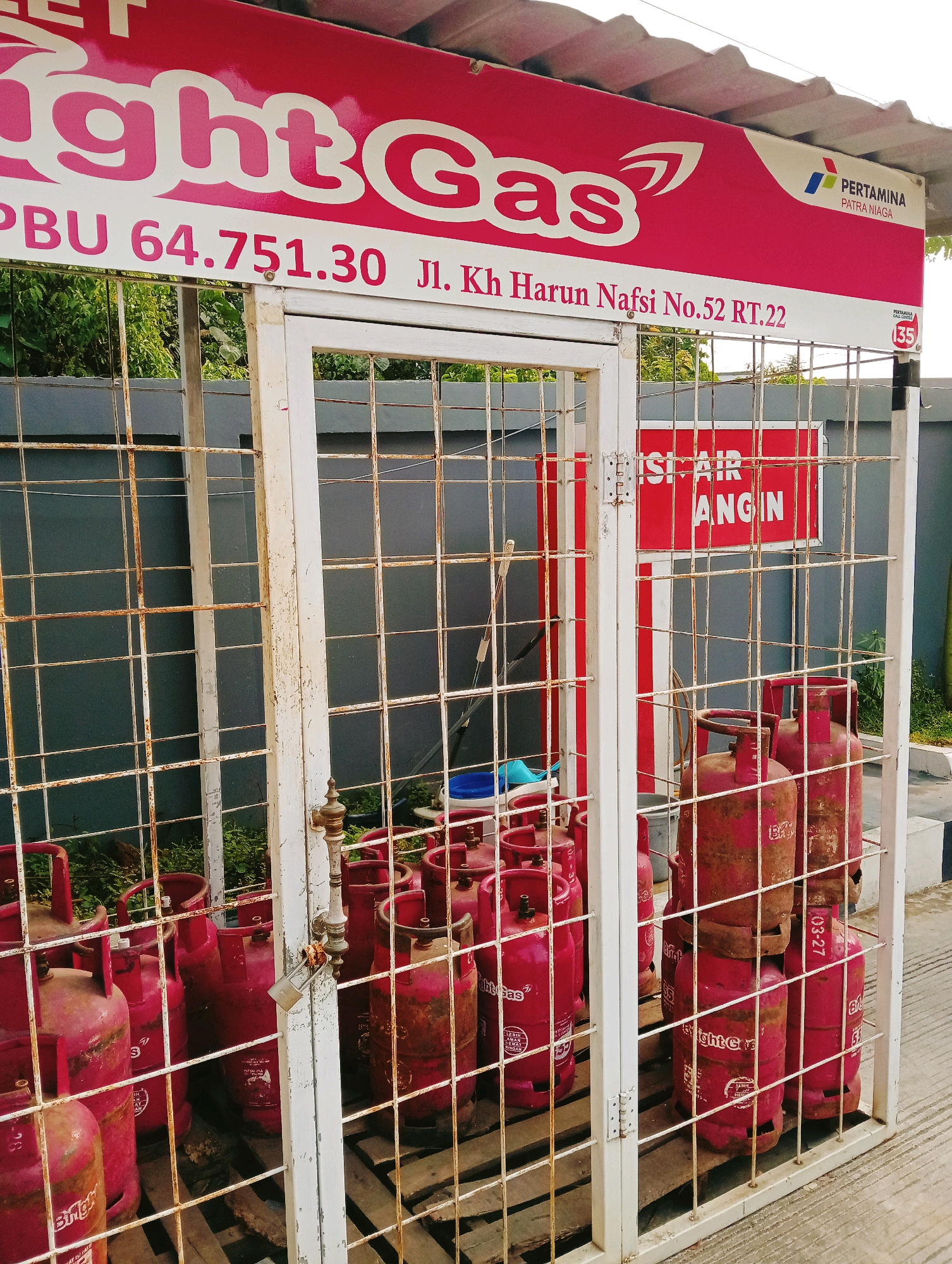 This is a close-up, eye-level, outdoor, daylight photograph of a propane gas cylinder storage area. The storage area is enclosed by a white metal cage with a padlock securing the entrance. The cage contains numerous red propane gas cylinders, some stacked on wooden pallets. A sign above the cage displays "IGHT GAS" in large pink letters, followed by "PBU 64.751.30 Jl. Kh Harun Nafsi No. 52 RT.22". Another sign on the right side of the frame shows "PERTAMINA PATRA NIAGA" and a smaller sign with "135". There is also a red sign inside the cage with white text that reads "ISI AIR ANGIN". The background shows a grey wall and green foliage. The scene appears to be a commercial establishment in Samarinda, Indonesia, likely a fuel station or a shop selling gas cylinders. There are no people visible in the image.