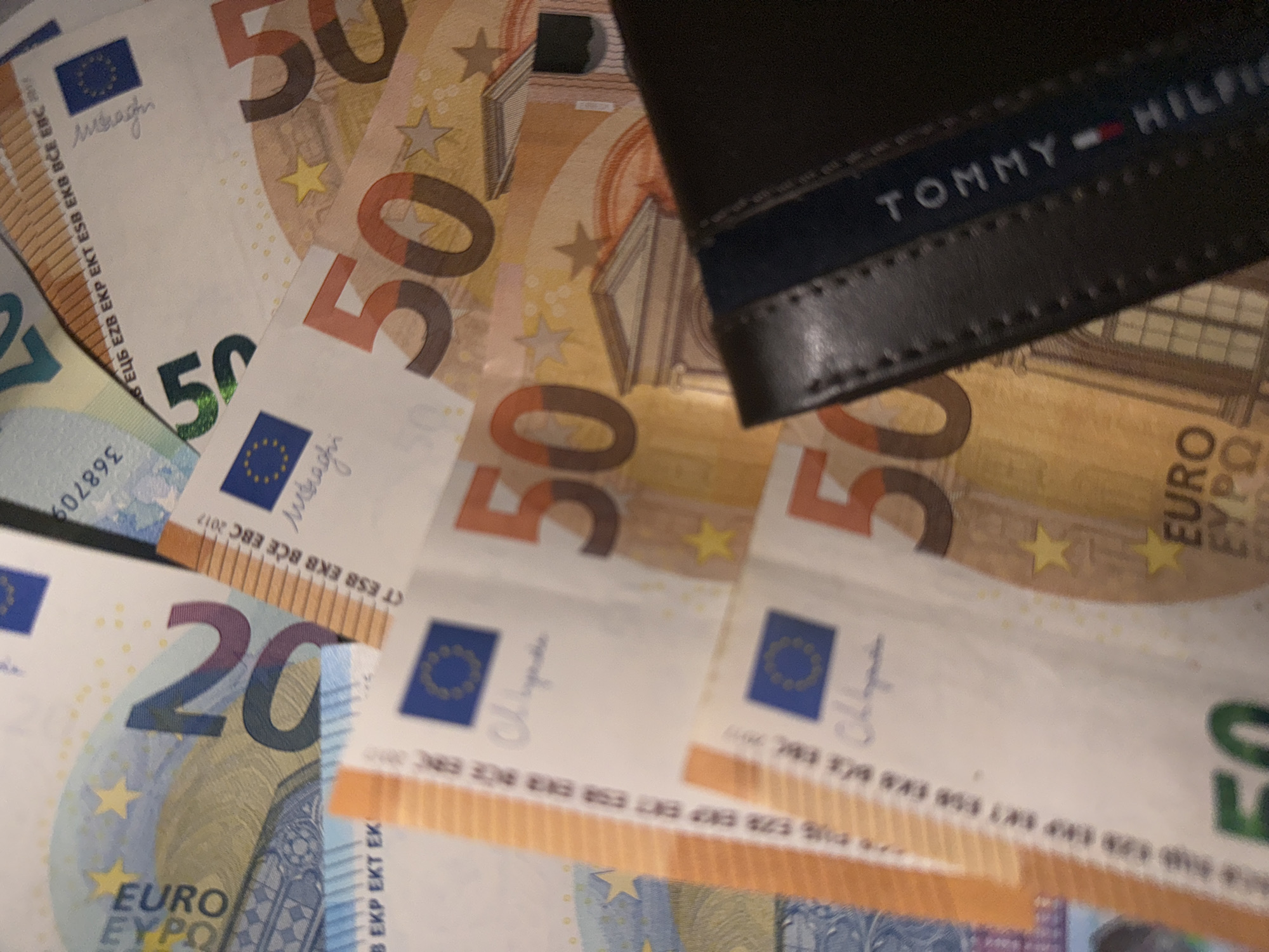 The image shows a close-up view of several euro banknotes of varying denominations, specifically 20 euro and 50 euro bills. The bills are scattered and partially overlapping. A dark-colored leather wallet with a "TOMMY HILFIGER" logo is placed on top of the banknotes.