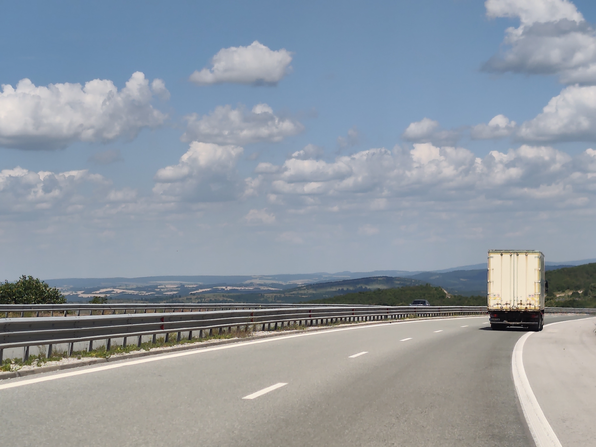 The image depicts a highway scene in Yablanitsa, Bulgaria. A light-colored truck with a closed trailer is traveling on the right lane of a paved road, curving to the right. The truck appears to be a delivery vehicle, and it is the main object in the foreground, with its rear end facing the camera. A dark-colored car is visible behind the truck in the distance, also on the highway.
The highway is bordered by a metal guardrail on the left, with grassy areas and some vegetation between the road and the guardrail. Beyond the guardrail, the landscape unfolds into rolling hills and fields, with some forested areas and a distant mountain range under a bright, cloudy sky. The terrain suggests a rural or semi-rural setting.
The time of day appears to be midday or early afternoon, indicated by the bright sunlight casting shadows and illuminating the scene. The weather is fair with scattered cumulus clouds in a blue sky, suggesting a pleasant day for travel.
There is no visible text in the image that provides specific information about the location or the subjects, apart from what can be inferred from the general landscape and the truck's design. The overall mood of the image is one of transit and open road travel.