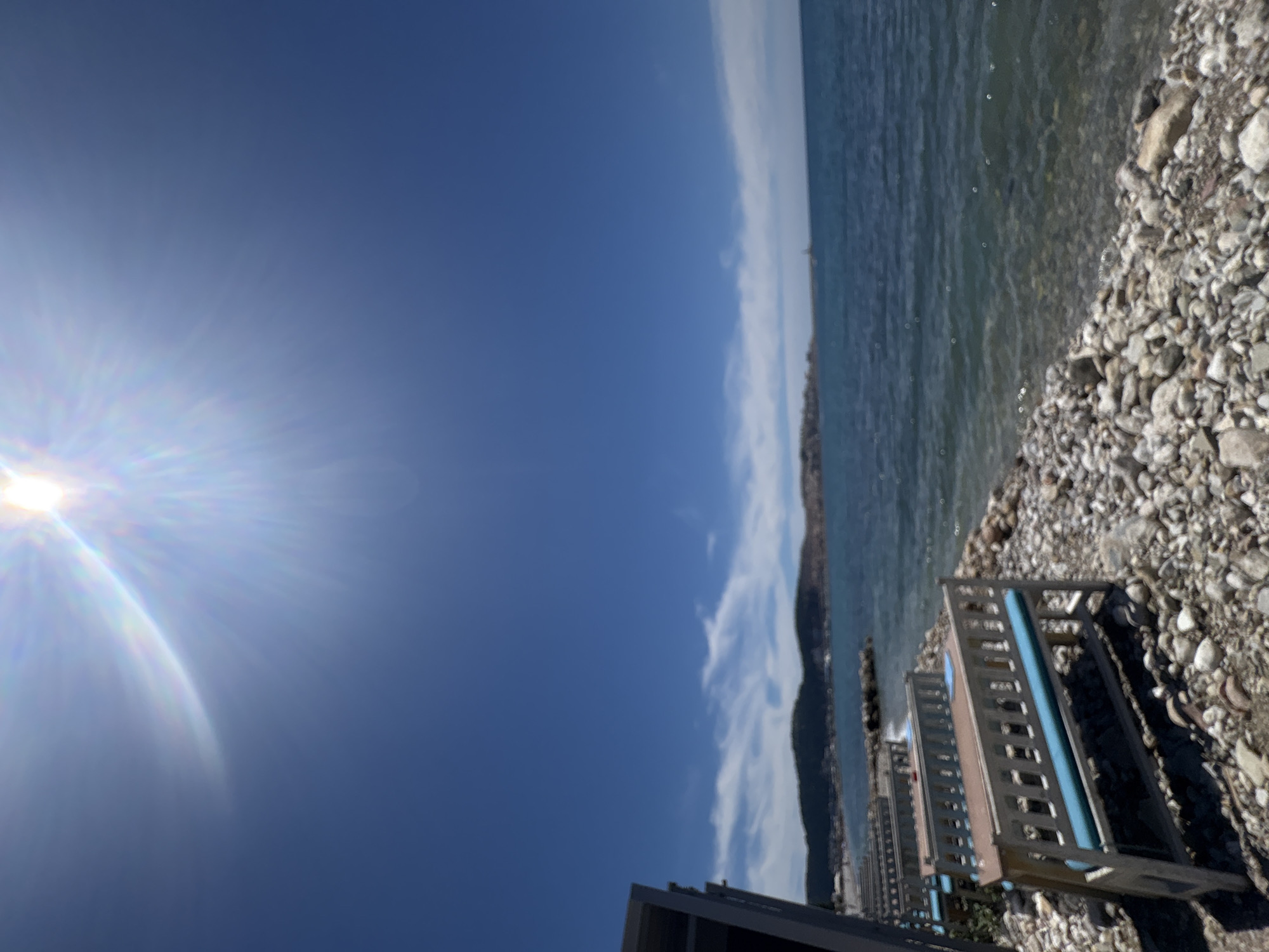 The image depicts an outdoor seating area on a pebble beach, likely in the city of Bar, Montenegro, overlooking a calm, blue sea under a bright, sunny sky. In the foreground, several empty wooden benches with bright blue cushions are arranged in rows. These benches appear to be part of a restaurant or cafe setup. The beach is composed of smooth, grey and white pebbles. The sea stretches out to the horizon, meeting a clear blue sky with a few wispy clouds. A distant, hazy coastline with hills and some structures is visible across the water. The sun is high in the sky, causing a lens flare and bright glare. There are no people or animals visible in the scene. The overall atmosphere is serene and suggests a pleasant, warm day.