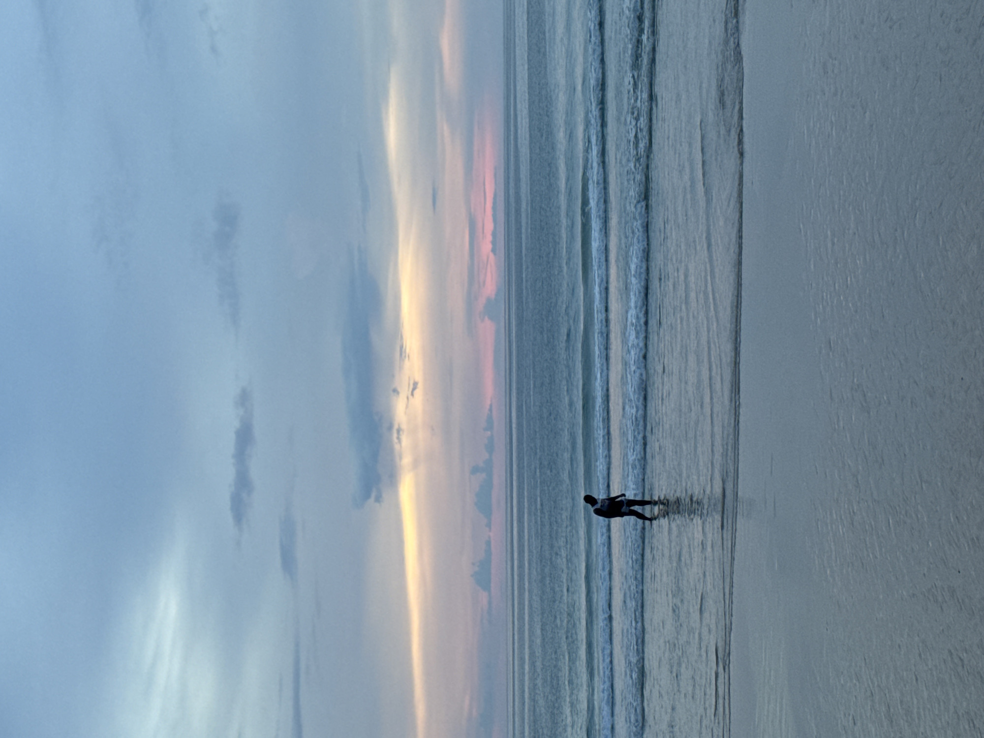 A person stands in shallow water on a beach in Choeng Thale, Thailand, facing left and looking down. The person is wearing a dark backpack. The sky above displays a gradient of colors from light blue at the top to pink and yellow near the horizon, suggesting a sunset or sunrise. The ocean has gentle waves breaking on the shore, and the wet sand reflects the sky.