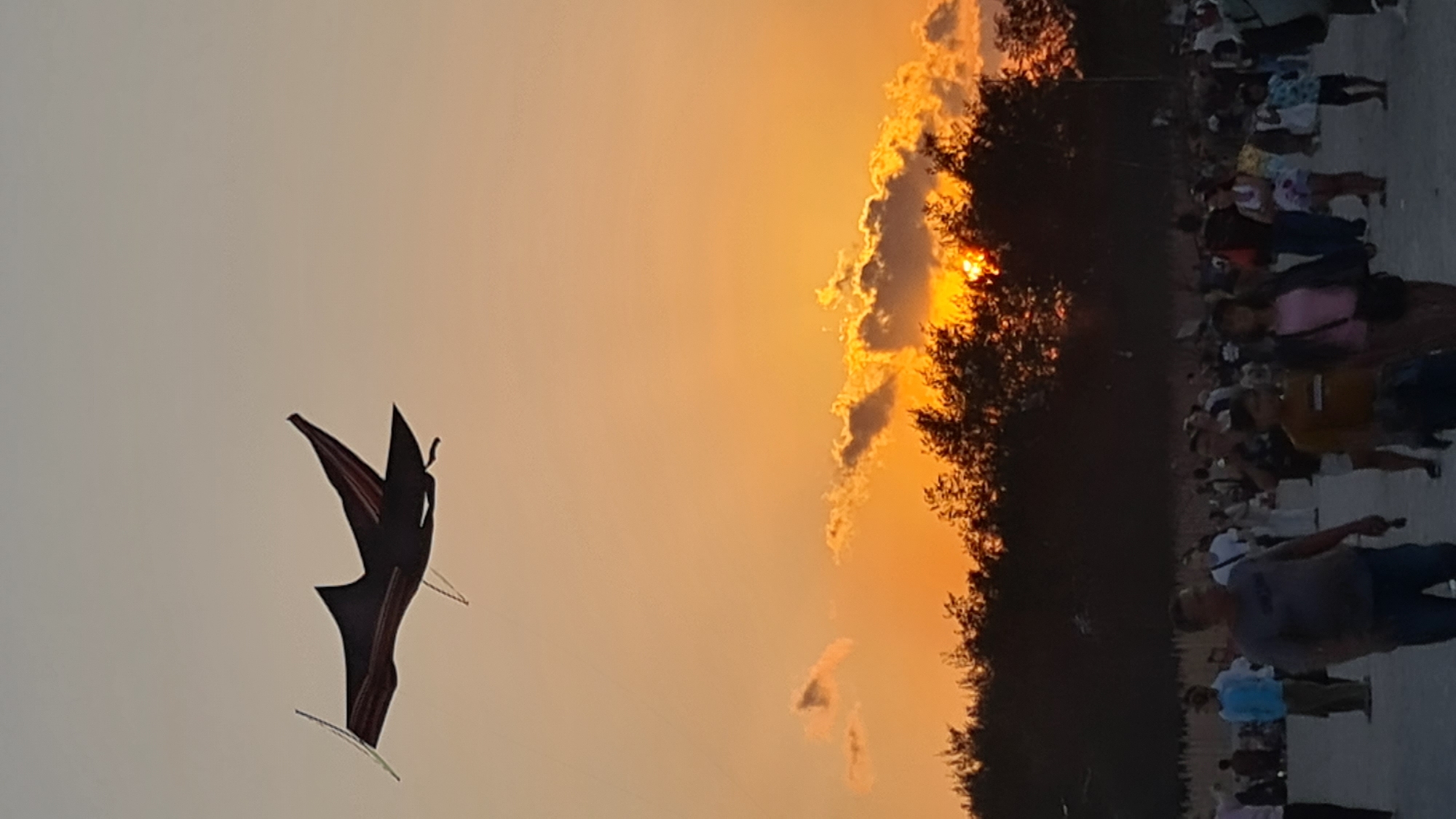 At sunset in Denpasar, Indonesia, a kite shaped like a shark flies against a sky illuminated by the setting sun. The kite, dark with red accents, is positioned in the upper left quadrant of the frame. Below, a silhouetted line of trees forms the horizon, with the sun peeking through the branches, casting warm orange and yellow light on scattered clouds. In the foreground, a crowd of people is gathered on a beach or open area, their figures silhouetted against the sunset. They are casually dressed and appear to be enjoying the evening. Some individuals are walking, while others are standing and observing the scene. The overall atmosphere is one of a relaxed, outdoor gathering during a vibrant sunset.