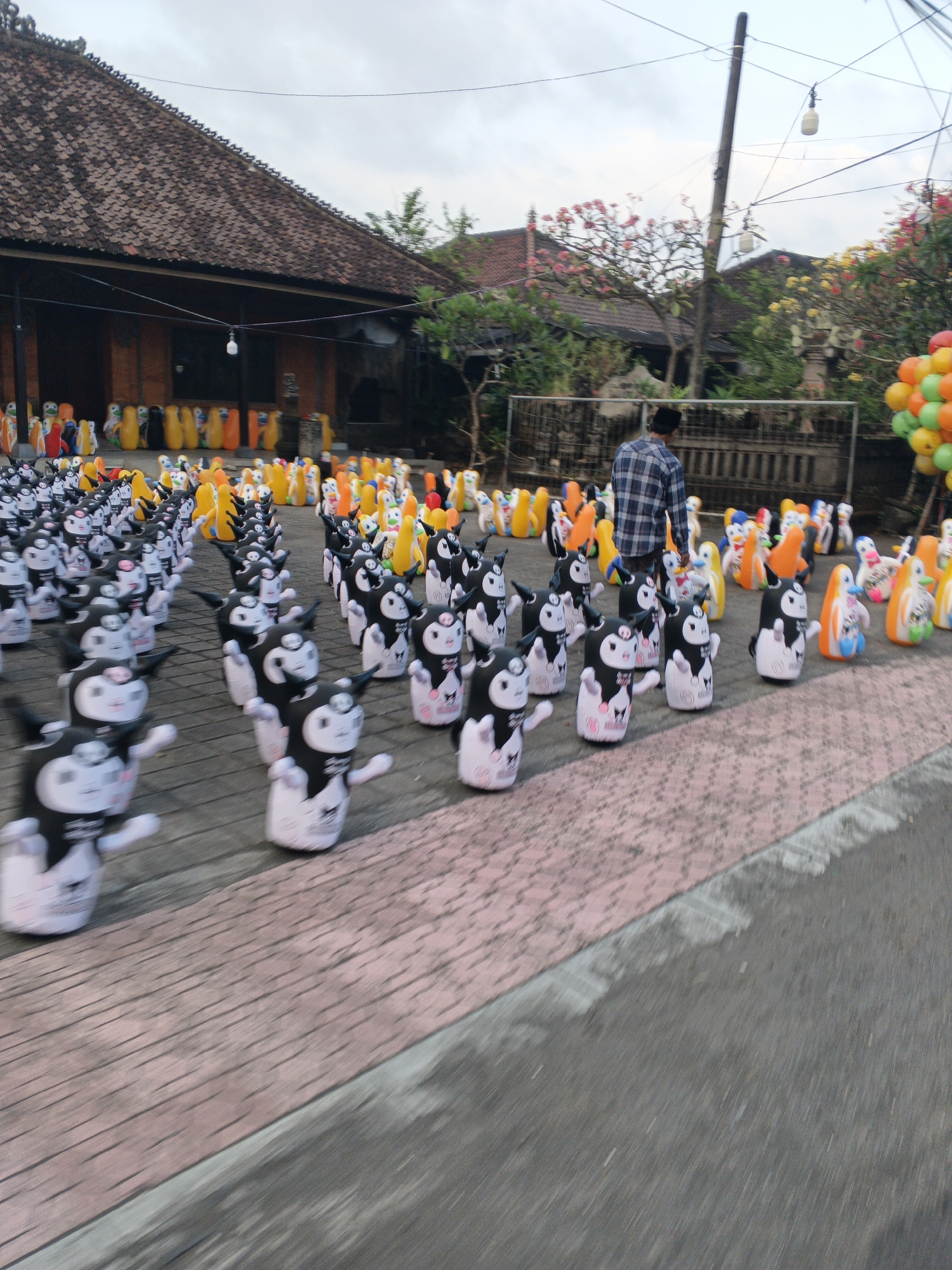 In Sukawati, Indonesia, an outdoor scene is captured with rows of inflatable cartoon figures lining a paved area, possibly a street or courtyard. The figures are predominantly black and white characters resembling penguins or similar cartoon animals, with some scattered yellow and orange inflatable shapes in the background. A man wearing a plaid shirt and a black peci hat is walking away from the camera, positioned between the rows of inflatables. Behind the figures, a traditional Balinese building with a tiled roof and wooden beams is visible. The sky is overcast, and there are power lines and hanging lights overhead. In the far right of the image, a bunch of colorful balloons is tied together. The overall impression is of an outdoor display or event, with the inflatables serving as decorations or attractions.