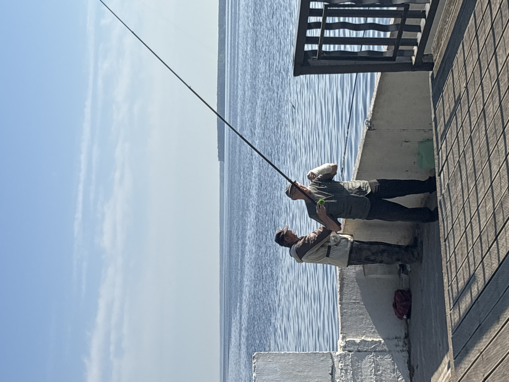 Two men are fishing from a pier in Chaikovsky, Russia. One man is holding a fishing pole while the other stands beside him. The pier has a wooden plank surface and a white cement wall. The sky is blue with some clouds. The water is calm with visible ripples.