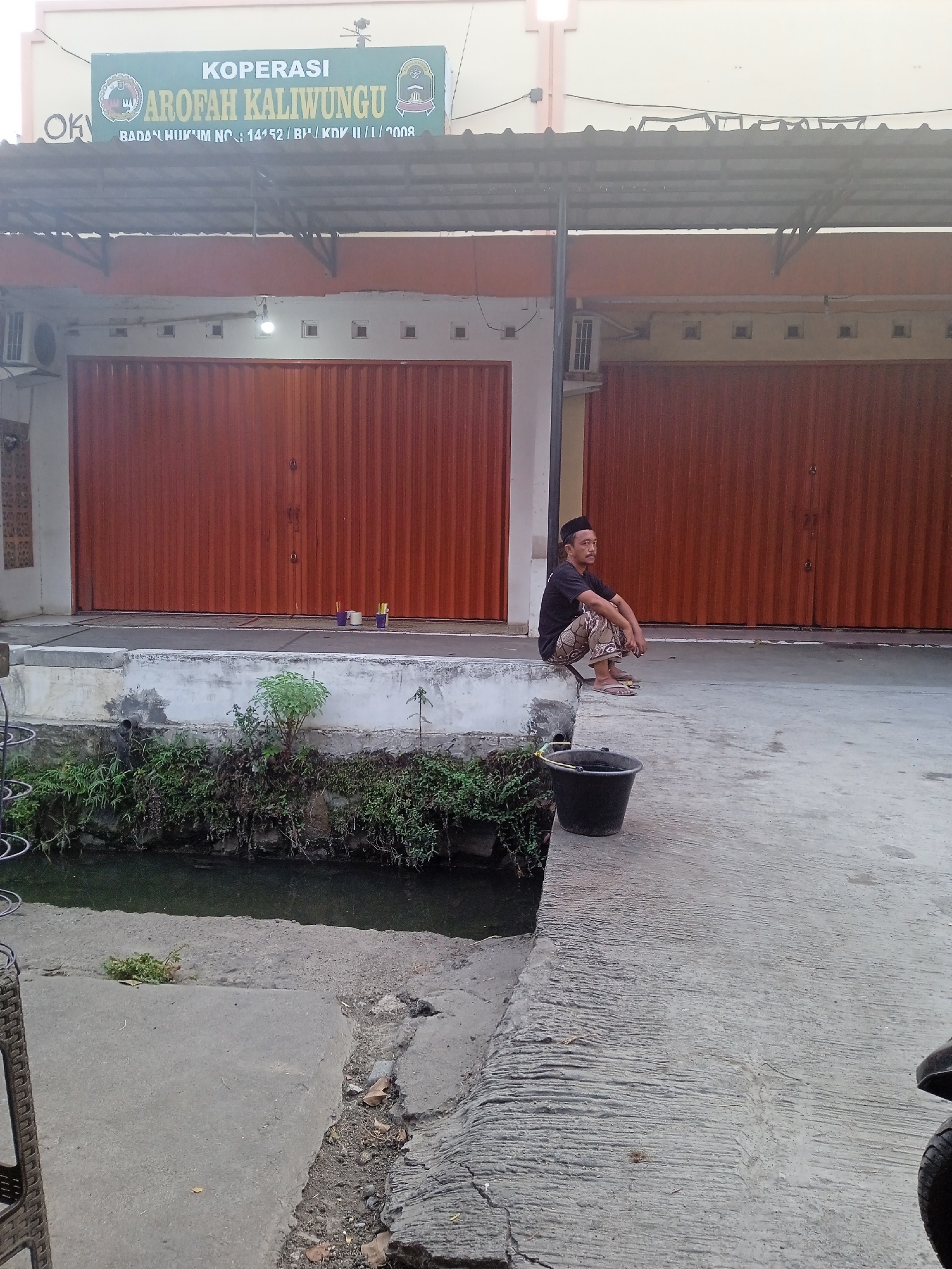 An Indonesian man sits on a concrete ledge next to a canal. He is wearing a black t-shirt, a black skullcap, and patterned pants. He has flip-flops on his feet. In front of him is a black bucket. The man is looking to his left. Behind him, there are two large, orange, roll-up doors that appear to be the entrances to shops. A sign above the doors reads "KOPERASI AROFAH KALIWUNGU" and also includes Indonesian text that translates to "Legal Entity No: 14152 / BH / KDK II / 1 / 2008." The shops appear to be in Sekopek Wetan, Indonesia. The canal is filled with murky water and surrounded by green foliage. The overall scene is a casual, everyday moment captured in an urban setting.