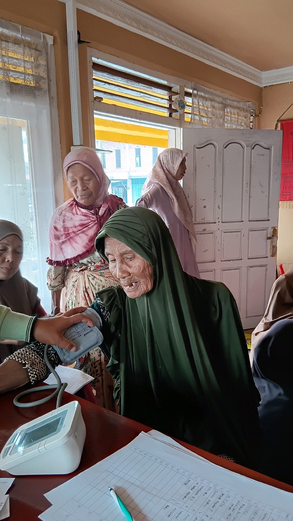 In the city of Padang Laweh, Indonesia, an elderly woman wearing a dark green hijab is having her blood pressure taken by an unseen individual. She is seated and her face, showing signs of age, is turned towards the blood pressure cuff attached to her arm. Behind her, two other women in hijabs are visible; one in a pink and patterned headscarf and dress, and another in a lighter colored hijab. A white digital blood pressure monitor and a pen lie on a red table in front of the seated woman, alongside a form with columns of text, suggesting a healthcare or administrative setting. The room has a window with blinds, and a door is visible in the background.