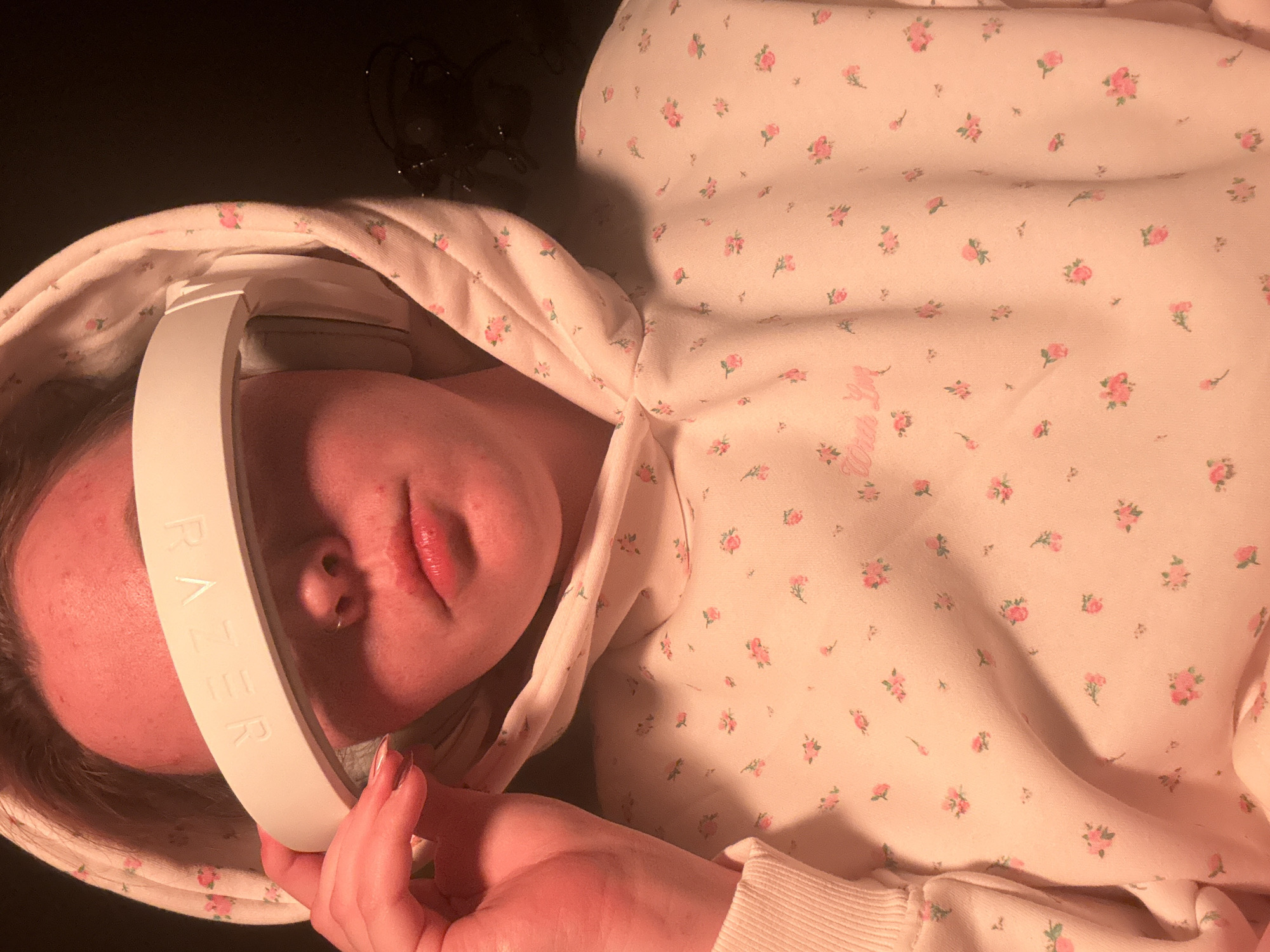 A person is lying on a dark surface, wearing a light pink hoodie with small floral prints, and holding a white Razer brand audio device over their eyes. The person's face is visible, showing their nose, lips, and forehead. A ring is in their nose. The location cannot be determined from the image.