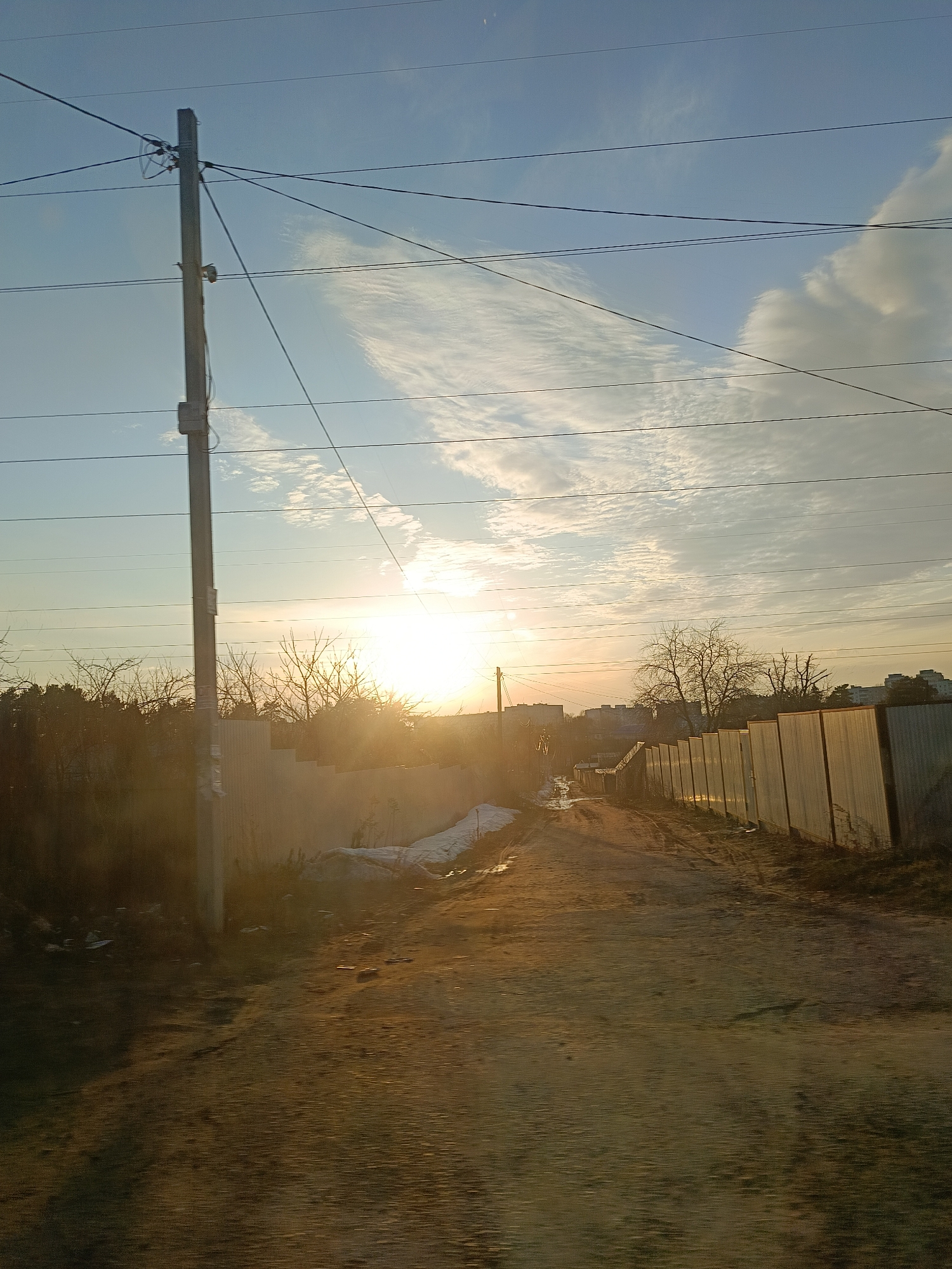 The image captures a dirt road flanked by fences in an urban setting. The sun is positioned at the end of the road, casting a golden light across the scene. Overhead are several power lines attached to a utility pole on the left side of the frame. Patches of snow remain near the base of the fence on the left side of the road. The sky is partly cloudy. Tall buildings are visible in the distance at the end of the road.