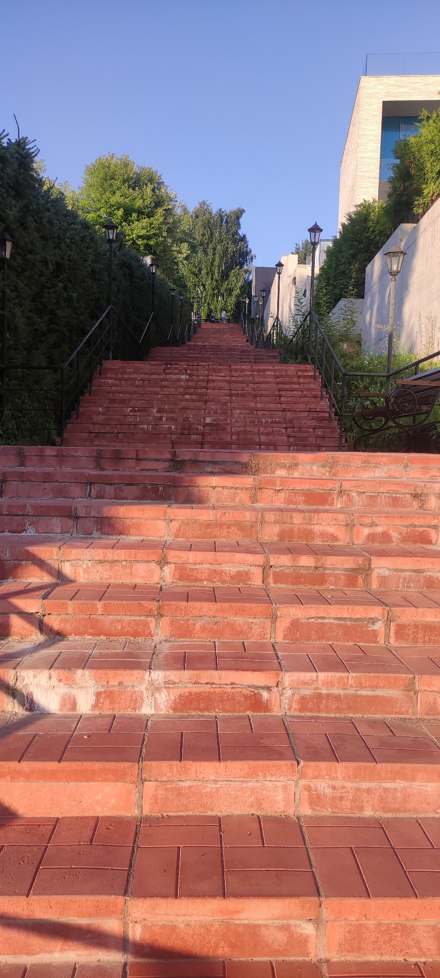 This is a vertical, eye-level, outdoor shot of a wide, red brick staircase leading upwards. The staircase is bordered on both sides by black metal railings and posts topped with black lamps. Lush green trees and hedges line the left side of the staircase, while on the right, there is a concrete wall and more greenery. Further up the stairs, on the right side, a dark metal bench with a wooden seat is visible. At the very top of the staircase, a modern white building with a blue-tinted window is partially visible, along with more greenery. The sky is a clear, bright blue, suggesting a sunny day. Two people are faintly visible at the top of the stairs, appearing to be walking. The scene is located in the city of Rosinka KP, Russia.