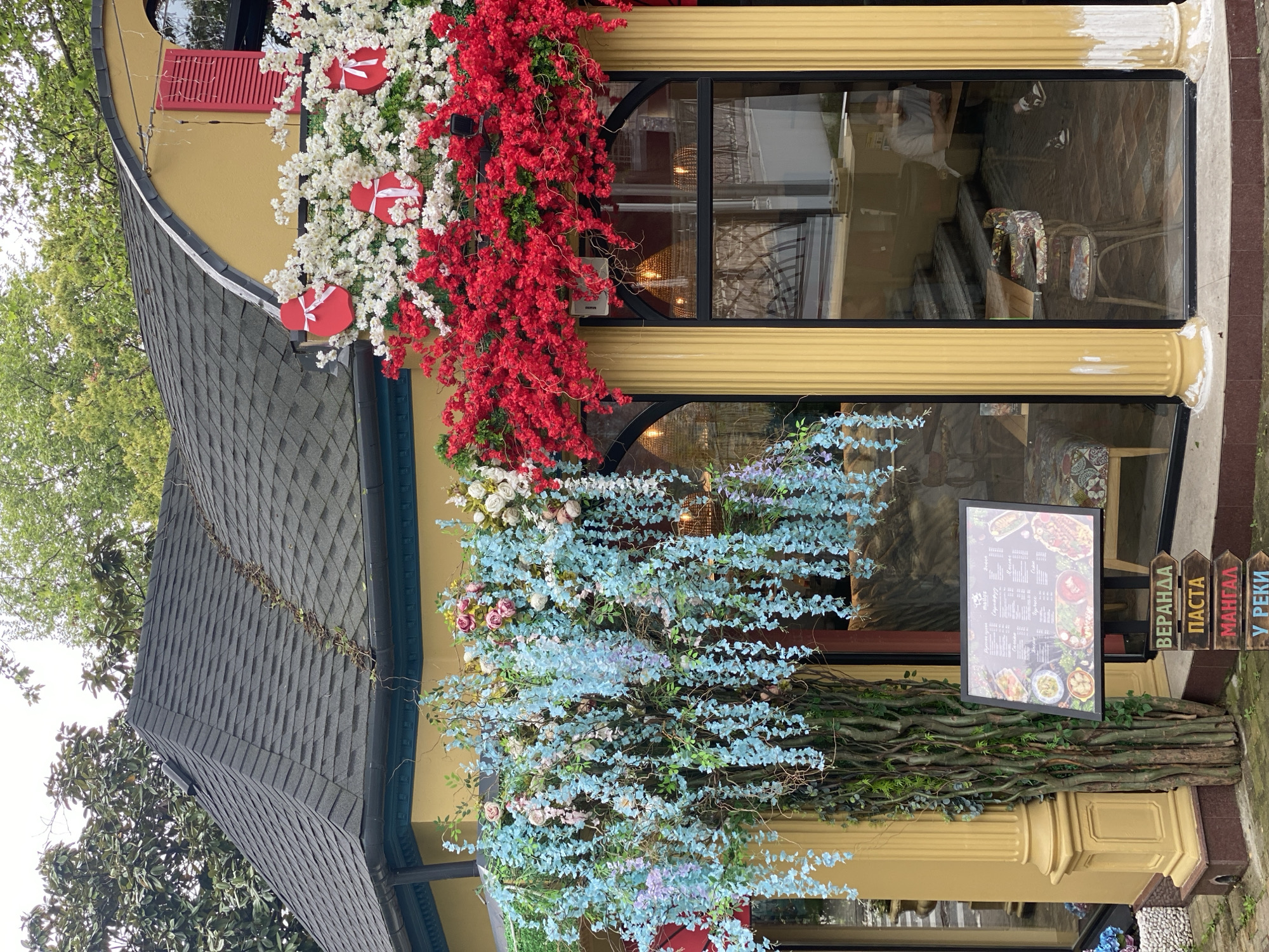 The image depicts the exterior of a building in Sochi, Russia, adorned with decorative floral arrangements. The building is yellow with black-framed windows, embellished with white and red flowers and heart-shaped accents near the top window, and blue and pink floral vines cascading below. A menu board stands near the entrance, along with wooden signs in Russian, indicating offerings such as "Veranda," "Pasta," "Mangal," and "U Reki." The architecture features a dark-gray, gabled roof, and the scene is partially framed by greenery, suggesting an urban or suburban setting.