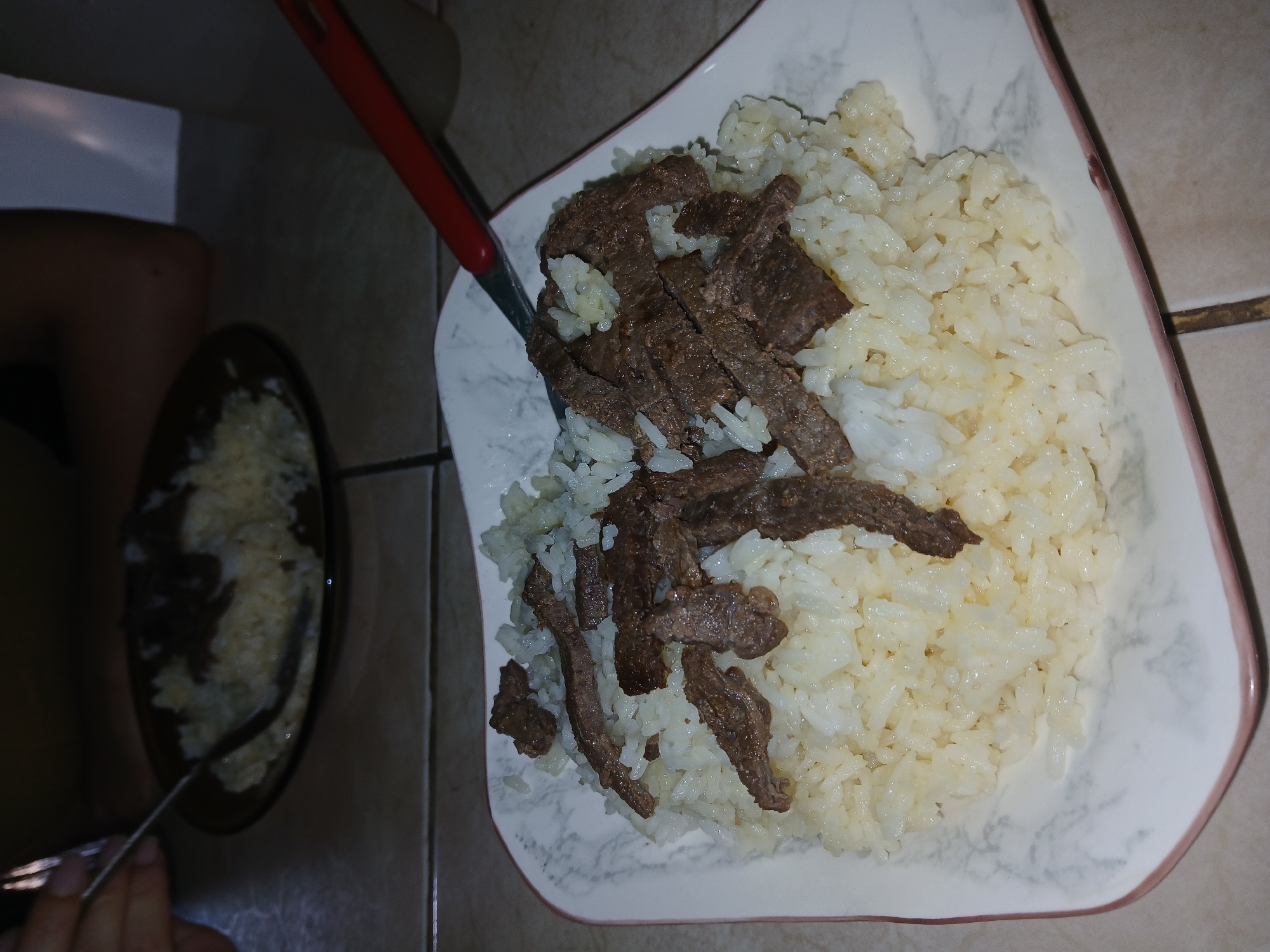 The image depicts a square white plate with a marble pattern and a pink rim, filled with rice and slices of cooked meat. A red-handled spatula rests on the plate. To the left, a similar dish of rice and meat sits on a dark surface. A hand holds a fork near the second dish. The background consists of beige tiled surfaces. The location is unspecified and cannot be determined to be Juan Griego, Venezuela from the image.
