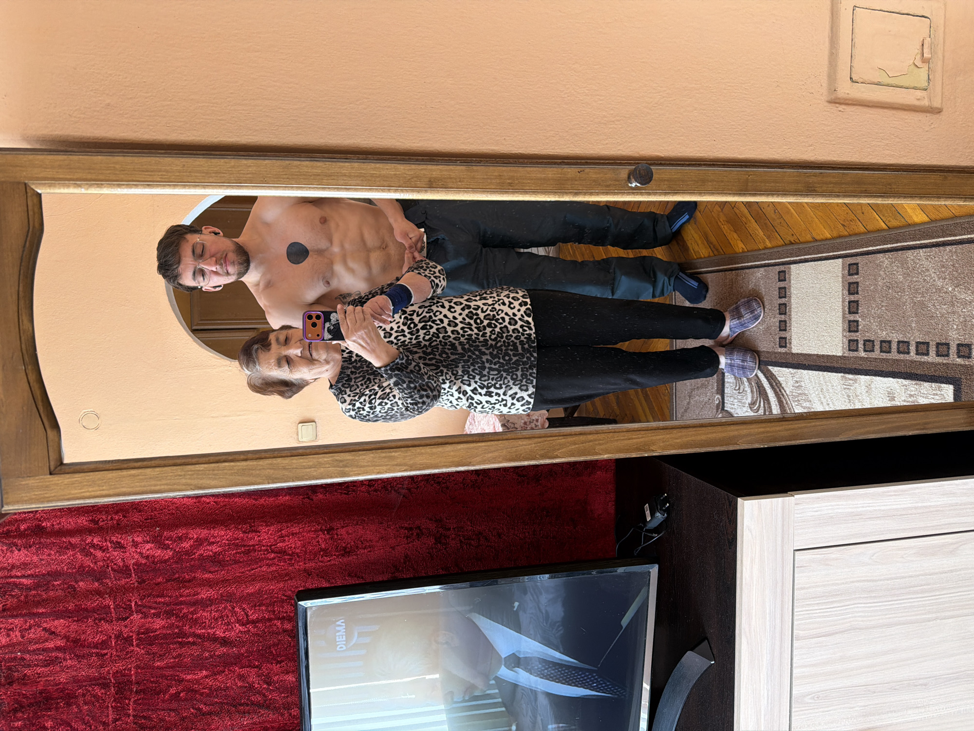 A mirror selfie shows a shirtless man with a black circular tattoo on his chest and glasses standing next to an older woman in a leopard print top and dark pants, who is holding a smartphone. Both are reflected in a large mirror. The background includes a peach-colored wall, a section of a rug with a geometric pattern, a red plush surface, and a television screen partially visible. The location appears to be indoors and the image implies it was taken at home. The city of Tsarevo, Bulgaria is not discernible from the image.