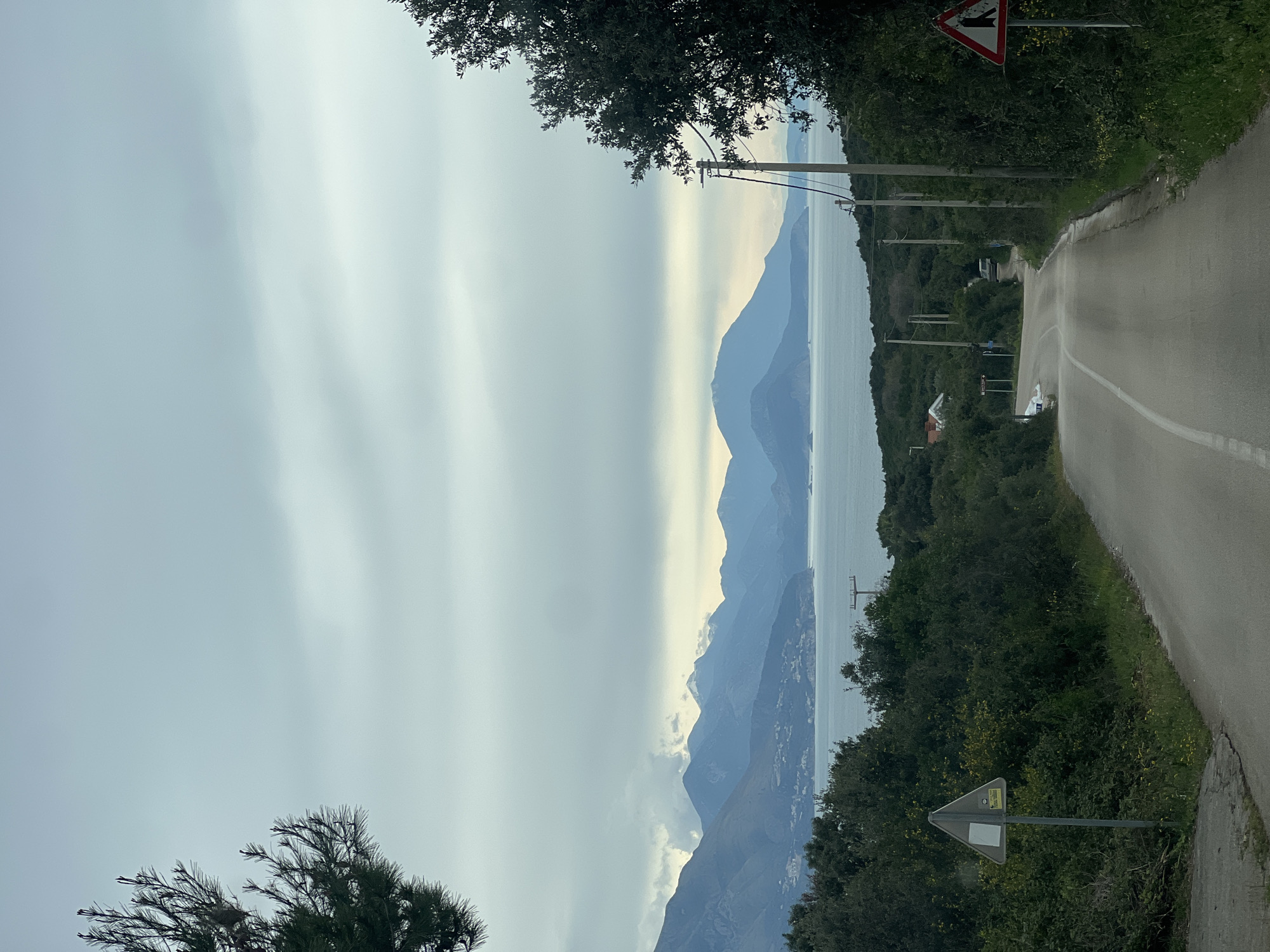 The image depicts a road in Krimovice, Montenegro, curving to the right with a white dividing line. The road is flanked by dense green vegetation and utility poles. In the distance, mountain ranges are visible across a body of water, under a cloudy sky. Two road signs are positioned near the roadside; one displaying an intersection symbol, and the other a generic triangular warning sign. No people are visible in the image.
