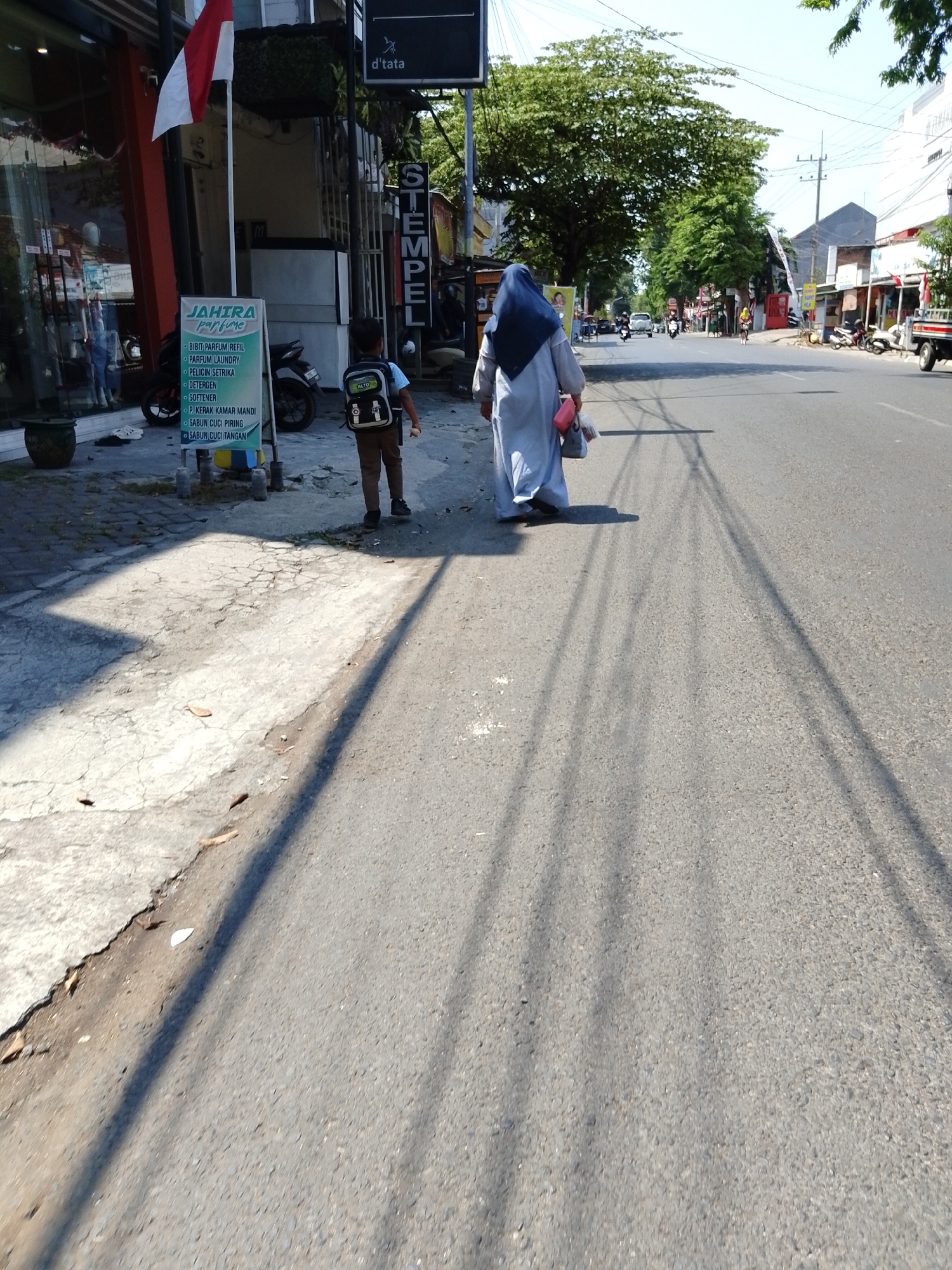A child wearing a backpack walks on the sidewalk of a street in Pasuruan, Indonesia. The child is facing away from the camera. To the right of the child, a person in a hijab and a light-colored dress walks on the street, also facing away from the camera. The person is carrying two bags. In the background, there are buildings, trees, and vehicles on the street, including motorbikes and a truck. The sidewalk is cracked and dry, with some leaves scattered on it. The street is paved and appears to be a typical urban street. The sky is clear and blue, indicating a sunny day. A sign in Indonesian is visible on the left, which appears to be an advertisement for a laundry service. The overall scene depicts a common street view in a city.