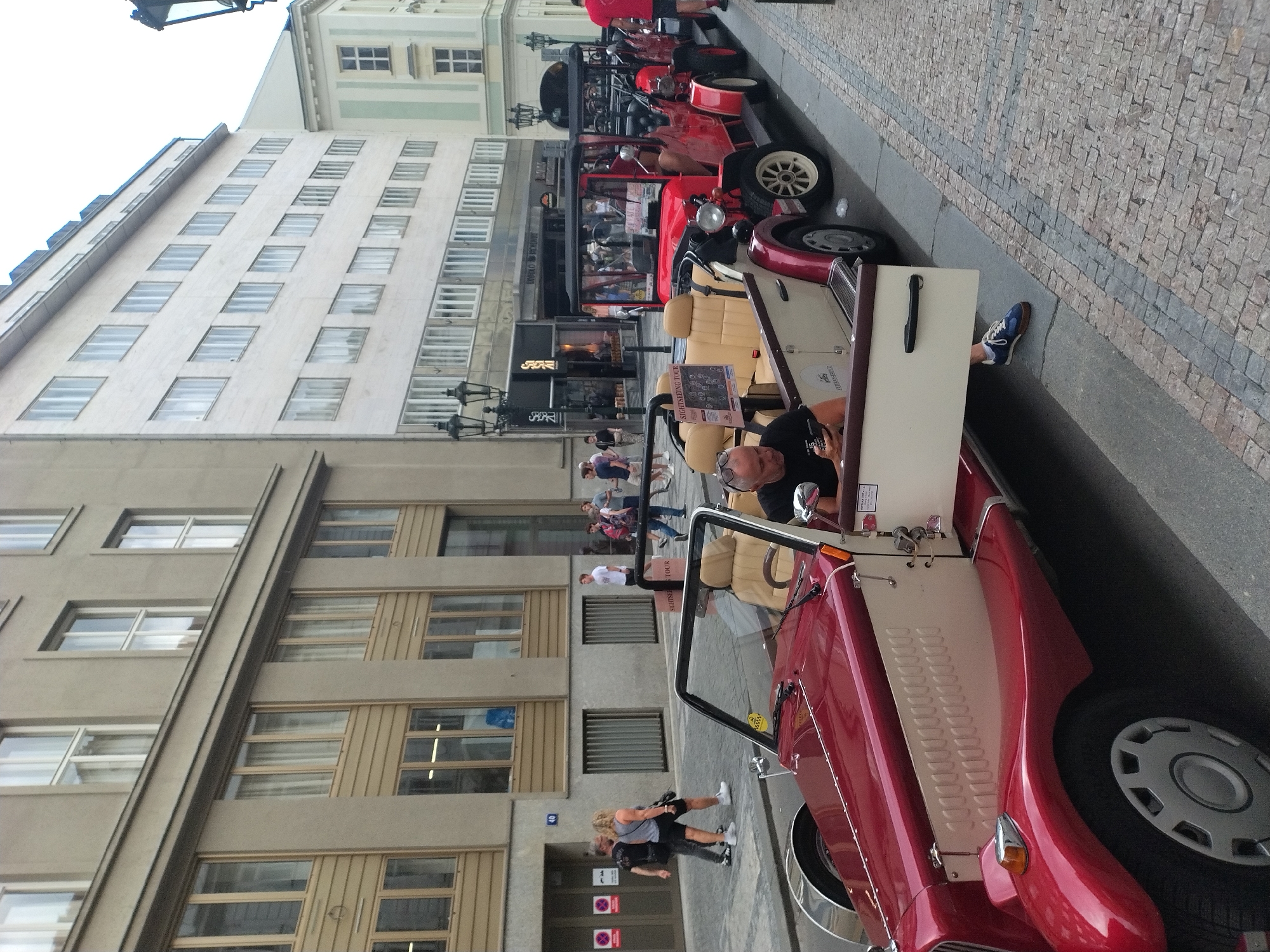 In Prague, Czechia, on a cobblestone street, a red and cream-colored vintage open-top car is parked. A man in a black t-shirt is seated inside the car, looking at his phone. Another vintage car, also red, is parked behind it. The street is lined with buildings, one of which has multiple floors with many windows. Several people are walking on the sidewalk in the background. One person is wearing a red shirt, and another is wearing a black shirt and dark shorts. The image captures a street scene in Prague, likely a tourist area, with vintage cars providing tours.