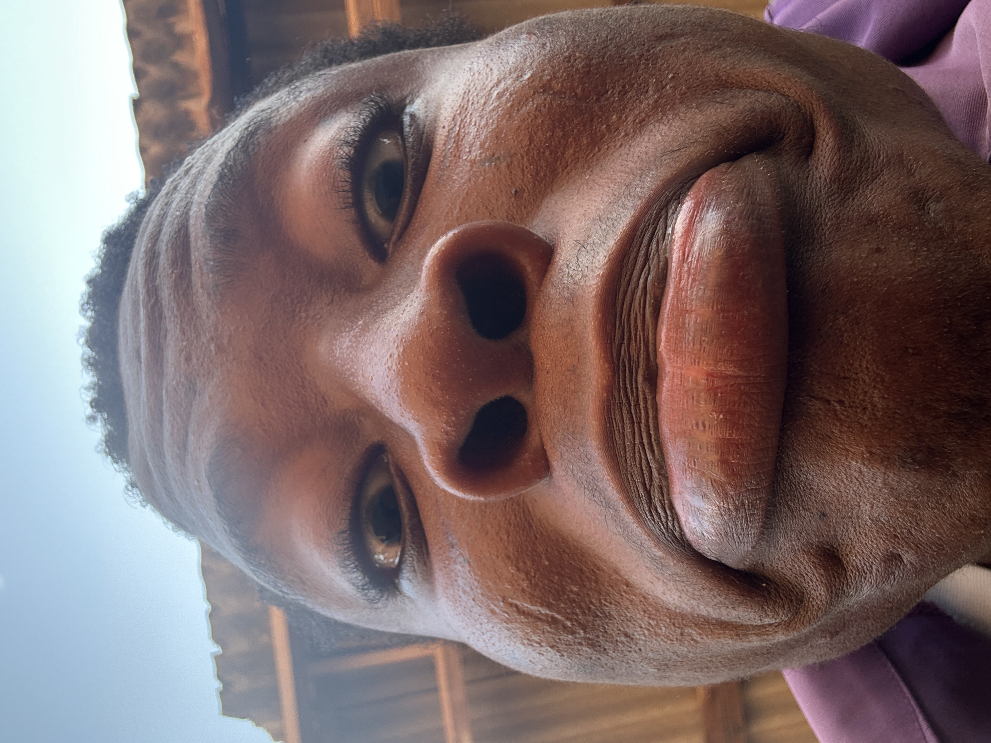 A close-up, low-angle selfie of a person with dark skin. The person is looking down at the camera, causing the facial features to appear compressed. The nose is prominent, and the lips are full. The eyes are partially open. The person has short, dark hair. A purple garment is visible on the right side of the frame. The background is blurred, but it appears to be an outdoor setting with wooden structures and a hint of sky in the upper left corner. Location: Unknown, Unknown.
