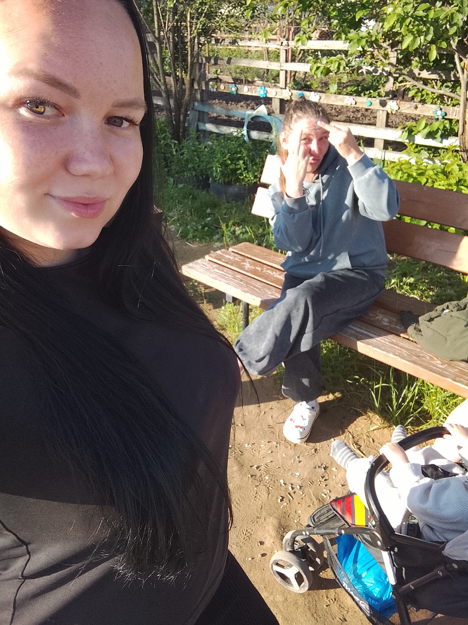 The image shows three individuals in an outdoor setting. A woman with dark hair and fair skin takes a selfie. In the background, a second woman sits on a wooden bench, holding tissues to her eyes. A child in a stroller is visible in the lower right corner. The ground appears to be dirt and sand. A wooden fence and greenery form the background. The setting appears to be in the city of Kuyeda, Russia.